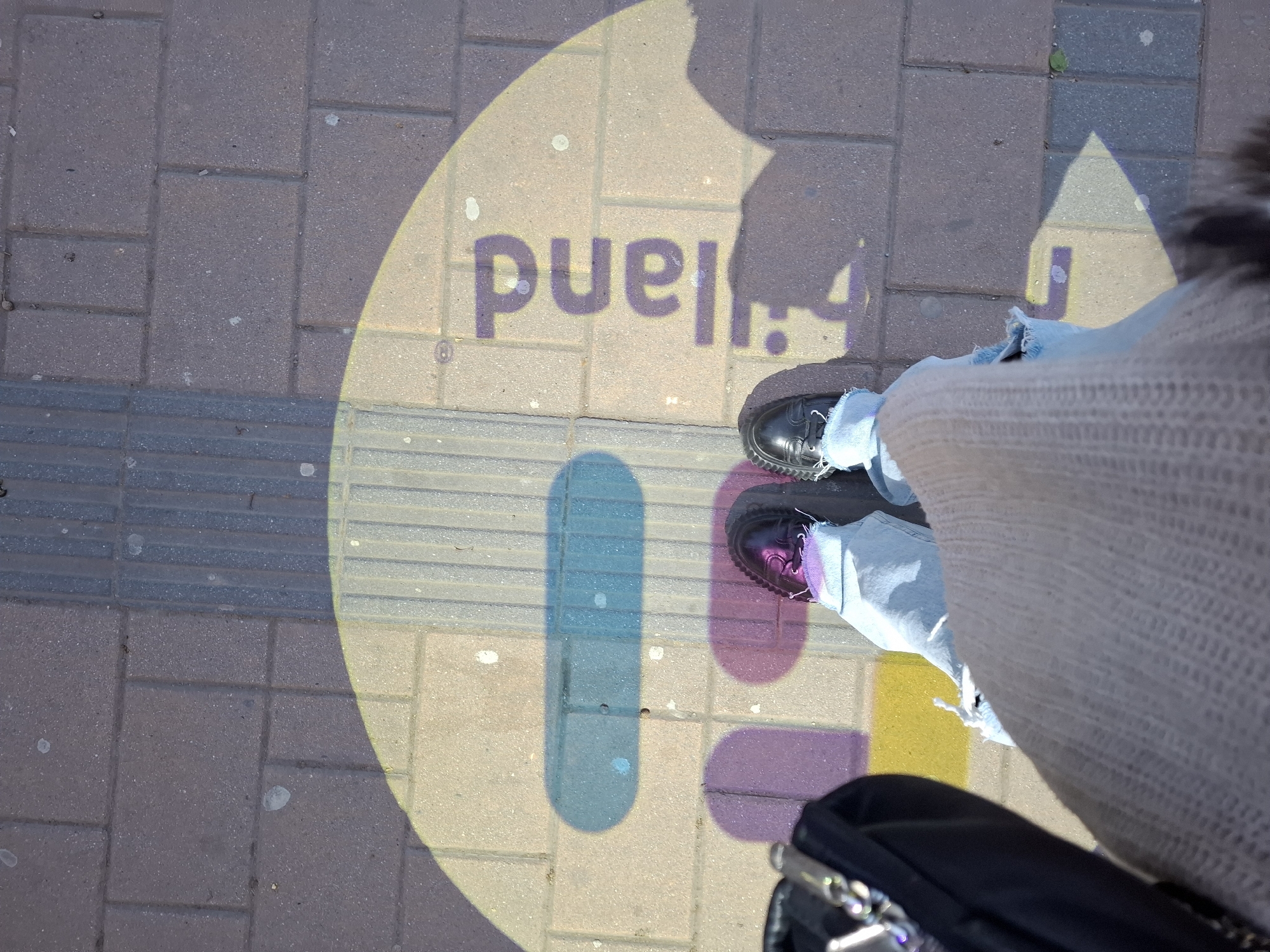 The image shows a person standing on a sidewalk made of rectangular paving stones in Novi Sad, Serbia. A shadow of the word "Fruška" is cast on the sidewalk, indicating the location is likely near Fruška Gora National Park. The person is wearing light blue jeans and black shoes with purple shoelaces. The bottom of a black bag or suitcase is visible near their feet. The sidewalk also has a section of tactile paving, typically used for visually impaired pedestrians. The overall lighting suggests it is daytime.