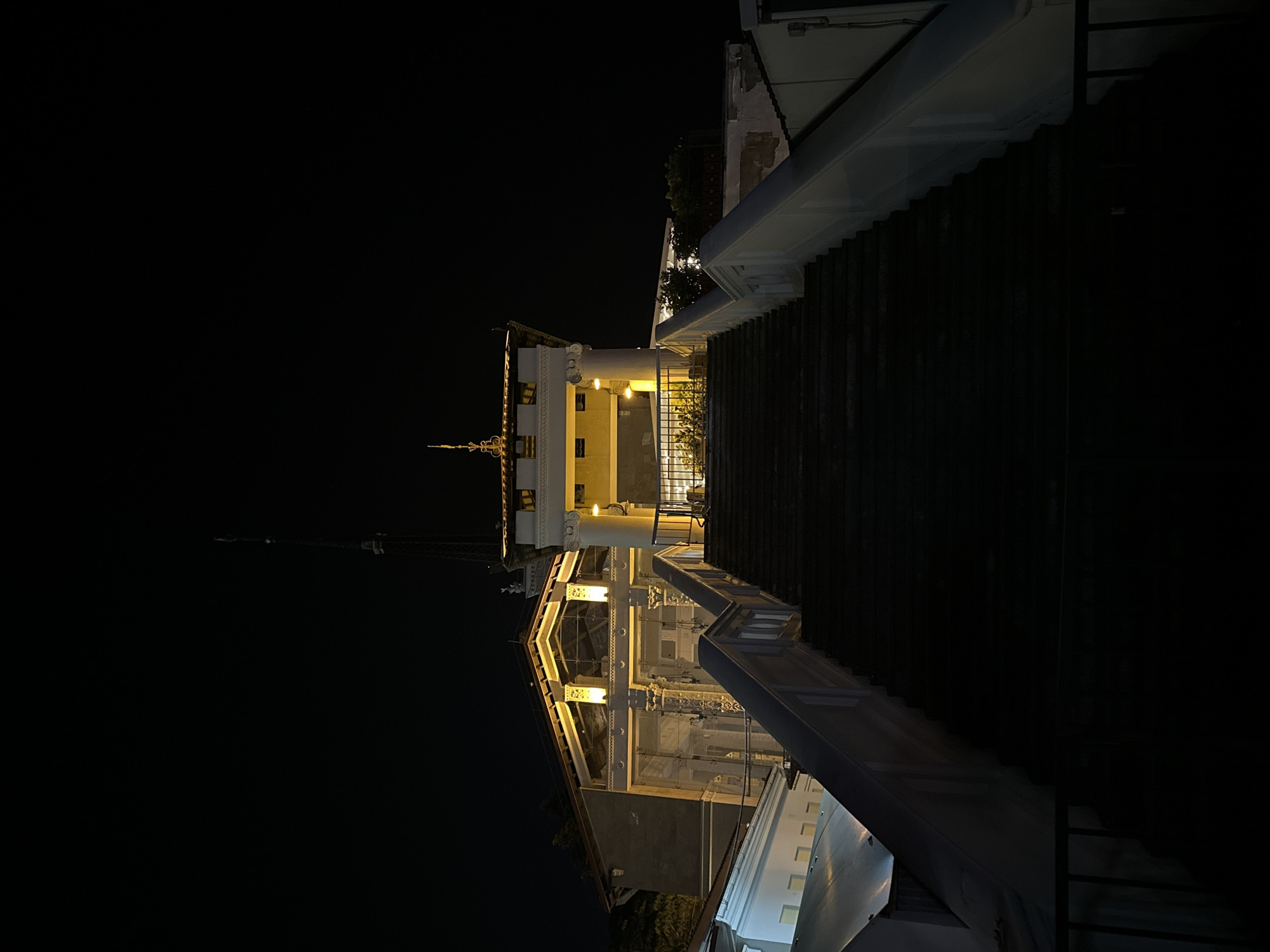 This is a low-angle, nighttime photograph of a grand staircase leading up to a building with classical architectural elements. The staircase is wide and dark, with white railings on either side. At the top of the stairs, the building features a portico with illuminated columns and decorative detailing. The facade of the building is a pale color, with yellow lighting emanating from within. A tall, thin tower, possibly a radio or communication tower, is visible in the background against the dark night sky. The overall impression is one of imposing architecture at night, with a focus on the ascending staircase. The specific location within the city of Sochi, Russia, is the Grand Hotel Zhemchuzhina.