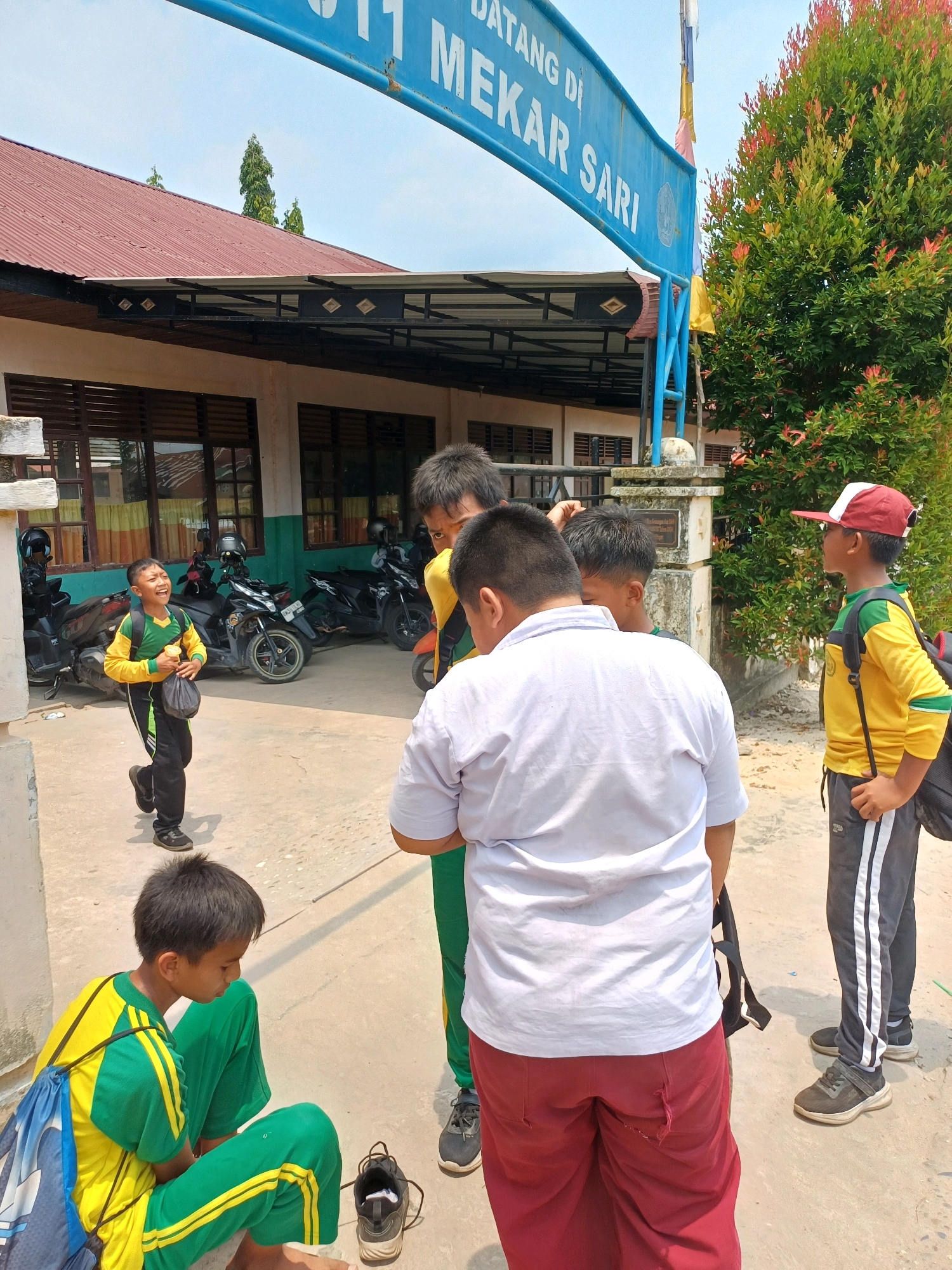 The image depicts a group of children, presumably students, gathered outside a building. The building appears to be a school, as indicated by the uniforms worn by some of the children and the presence of motorcycles, likely belonging to staff or students. A blue sign above the entrance reads "DATANG DI MEKAR SARI," suggesting the location is a school named Mekar Sari in Dumai City, Indonesia.
Several children are visible, dressed in yellow and green uniforms, which are common in Indonesian schools. One boy in a yellow and green uniform is running and laughing, holding a plastic bottle and a bag. Another child sits on the ground, also in a yellow and green uniform, with a blue backpack beside him. Two boys in the foreground are facing away from the camera, one wearing a white shirt and red pants, the other in a yellow and green uniform with a red and white cap. A boy in a yellow shirt and dark pants with white stripes is standing on the right, looking to the side.
The environment is outdoors, with a concrete ground and a building with a red corrugated metal roof. There are several motorcycles parked in front of the building. A large green tree with red-tinged leaves is visible to the right of the entrance. The sky is clear and blue. The overall impression is of a school setting during a break or at the end of the school day.