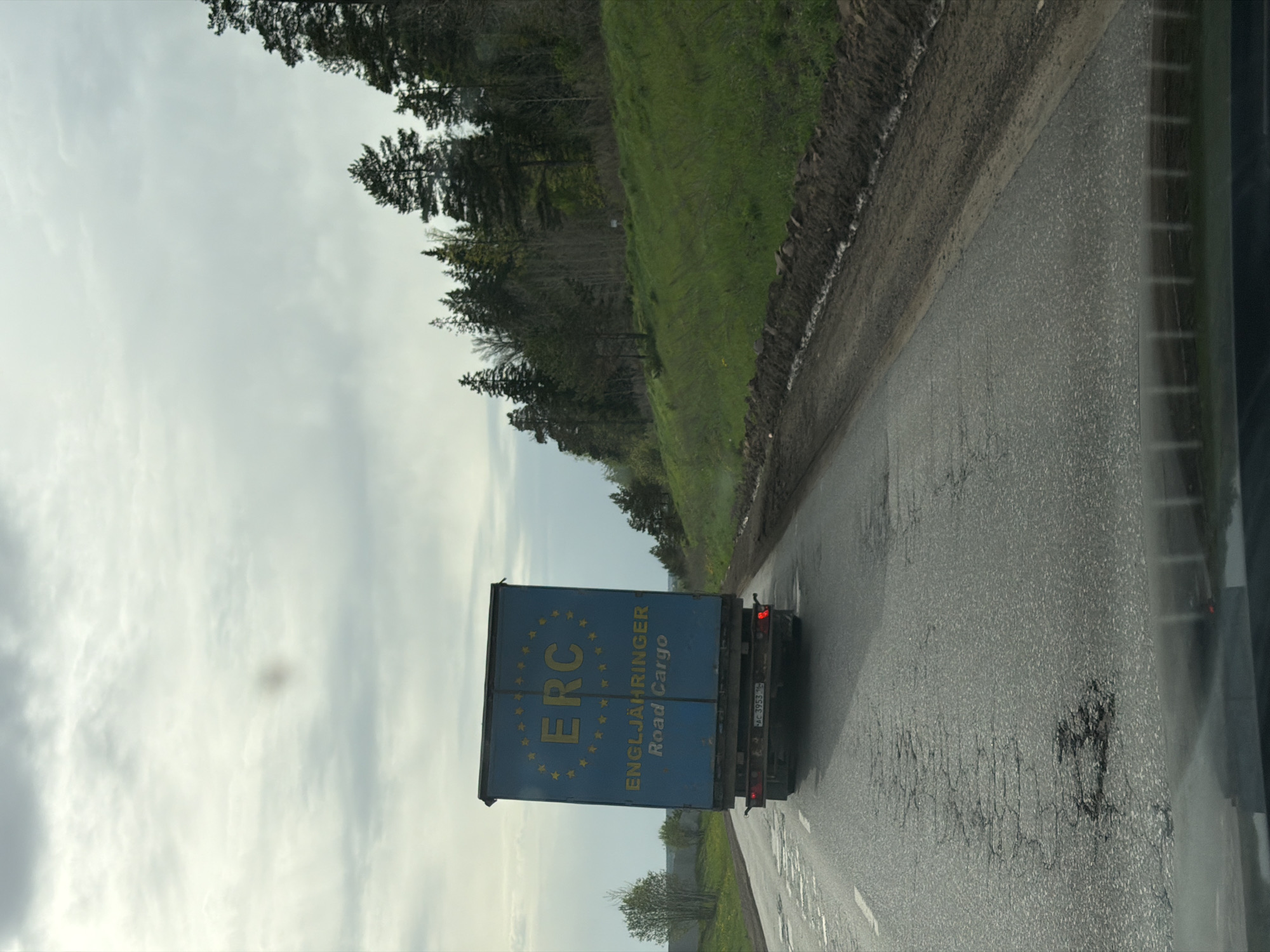 The image depicts a blue delivery truck on a road in Staryi Chul'tem, Russia. The truck has yellow lettering on its side that reads "ERC ENGLJAHRINGER Road Cargo." The sky is cloudy and there are trees lining the side of the road. The road surface appears wet. The image was likely taken from a vehicle moving behind the truck.