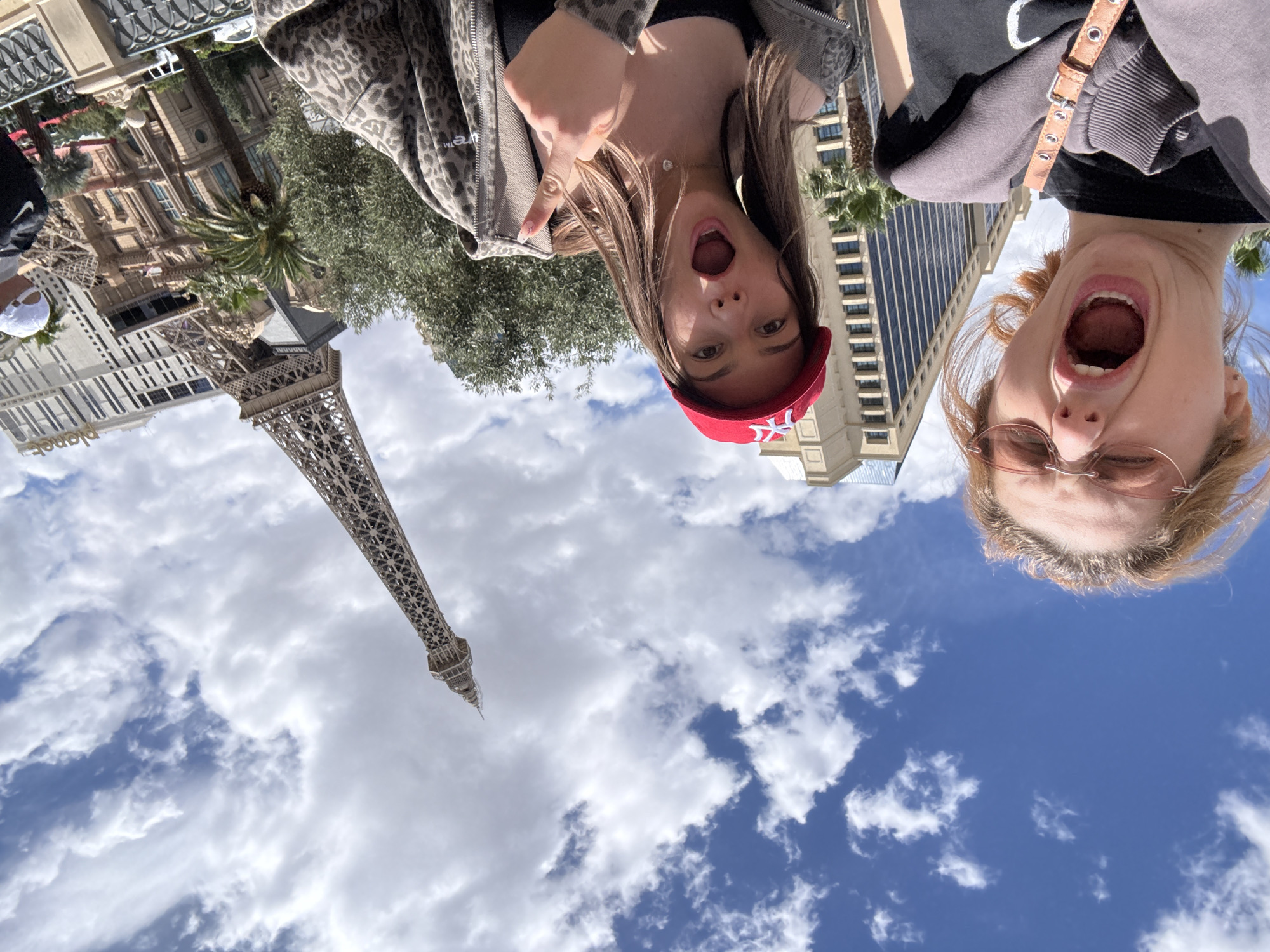 The image shows two women in Paradise, Nevada, United States, posing with their mouths open in front of a view of the Eiffel Tower replica at the Paris Las Vegas Hotel & Casino. The taller woman is on the left, wearing a red baseball cap and leopard print jacket, and is pointing. The shorter woman is on the right, wearing glasses and a gray jacket. The background includes the tower, building facades, clouds, and sky.