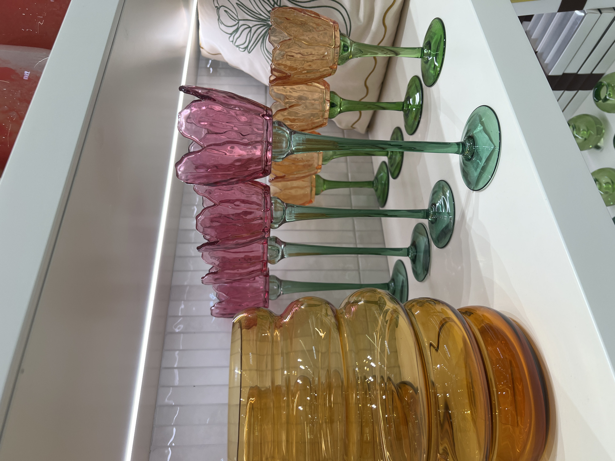 The image depicts a retail display of glass home decor items on a white shelf, likely in a shop in Saint Petersburg, Russia. In the foreground, a large, fluted amber glass vase is positioned on the left. To its right, a row of decorative glass candle holders with tulip-shaped tops are arranged. These candle holders feature pink tops and green stems, with some appearing to be in amber and green as well. The background shows a white tiled wall and the edge of a shelf above, illuminated by strip lighting. A decorative pillow with a green line drawing of a plant is partially visible behind the candle holders on the right. The overall scene suggests a well-lit retail environment showcasing aesthetic glassware.