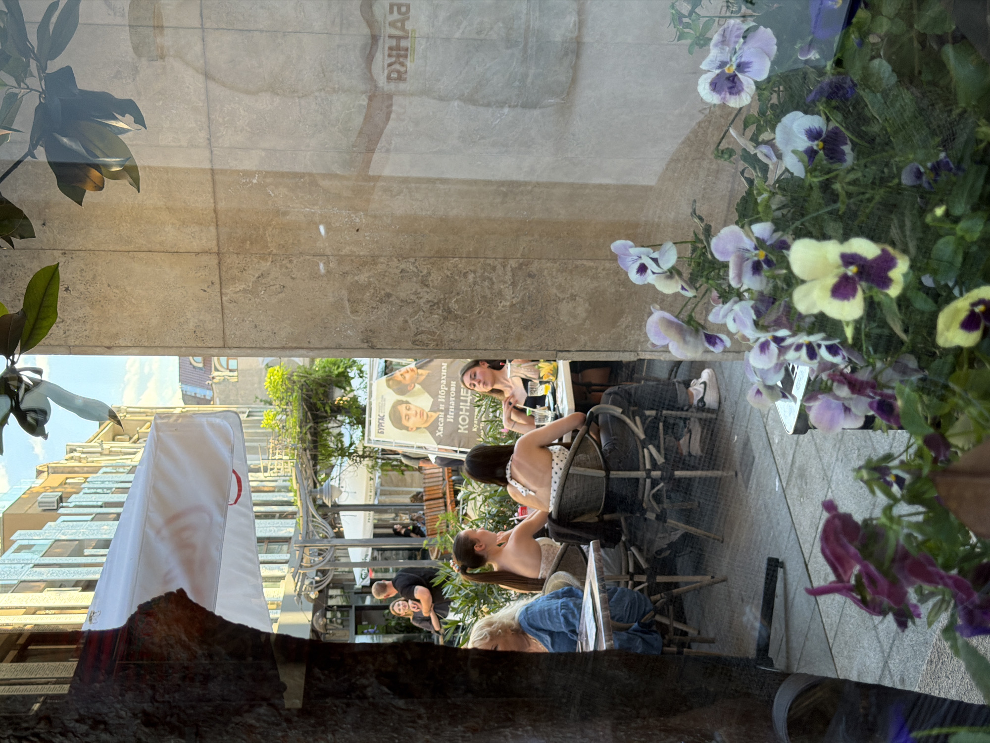 The image shows an outdoor cafe scene in Burgas, Bulgaria. In the foreground, a reflection of green leaves and purple and yellow pansies is visible on a glass surface. Beyond the reflection, people are seated at tables under a white umbrella. A poster for a concert featuring "Hasan and Ibrahim Ignatov" is displayed on an easel. In the background, buildings line a street, suggesting an urban environment. The overall ambiance is that of a casual outdoor gathering.