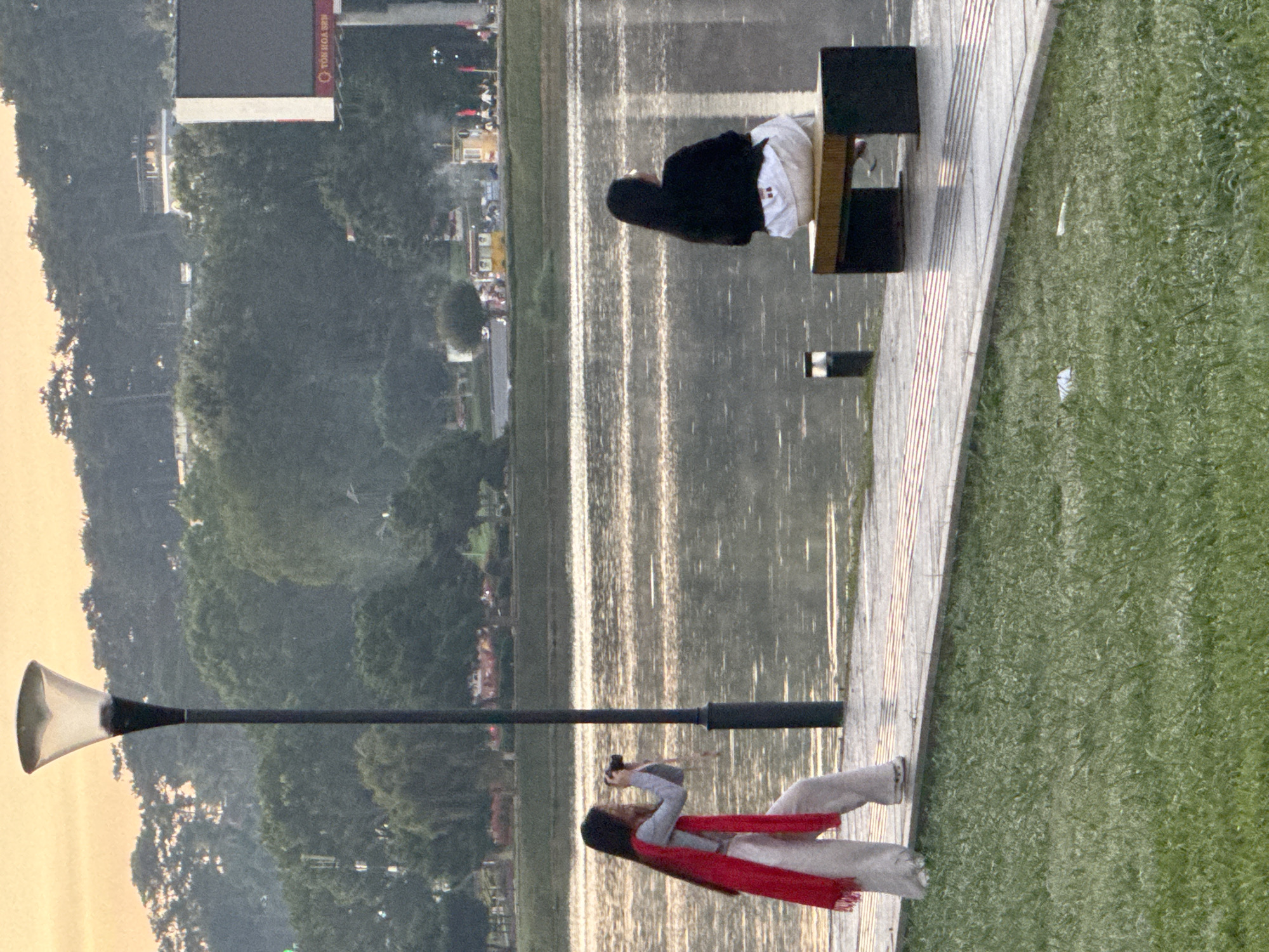 A woman wearing a red scarf and white pants photographs a person sitting on a bench by Xuan Huong Lake in Da Lat, Vietnam. The woman is positioned on a paved walkway next to a grassy area, while the seated person faces away from the camera, looking towards the lake. A street lamp is visible to the left of the frame, and trees and hills form the background. The sky is overcast with a warm hue, suggesting either dawn or dusk.