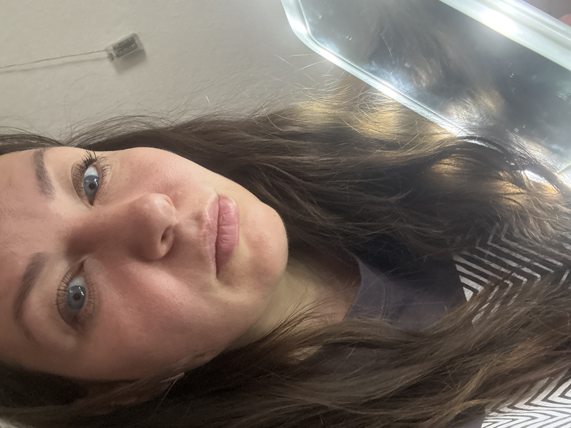 An overhead portrait features a young woman with a neutral expression, positioned with her head tilted towards the left side of the frame. She has blue eyes, dark eyelashes, and well-defined eyebrows. Her light skin tone and full lips are visible. Long, wavy dark brown hair cascades around her head and shoulders, with hints of lighter brown or highlights.
The setting appears to be indoors, likely a room with a light-colored ceiling. In the upper left background, a small, light-colored electronic device, possibly a sensor or speaker, with thin wires is mounted on the ceiling. In the upper right quadrant, a reflective, rectangular object, possibly a mirror or a screen, reflects some light and a distorted view of the surroundings or hair.
The woman is wearing a dark-colored garment, visible near her neck. Further to the right, under her hair, a fabric with a black and white geometric pattern (zigzags) is visible, suggesting bedding or another piece of clothing. The lighting appears to be ambient indoor light, possibly from above. The image captures a serene moment, with no specific activity indicated other than posing for the photograph. There is no visible text in the image. The implied location context is Chaikovsky, Russia.