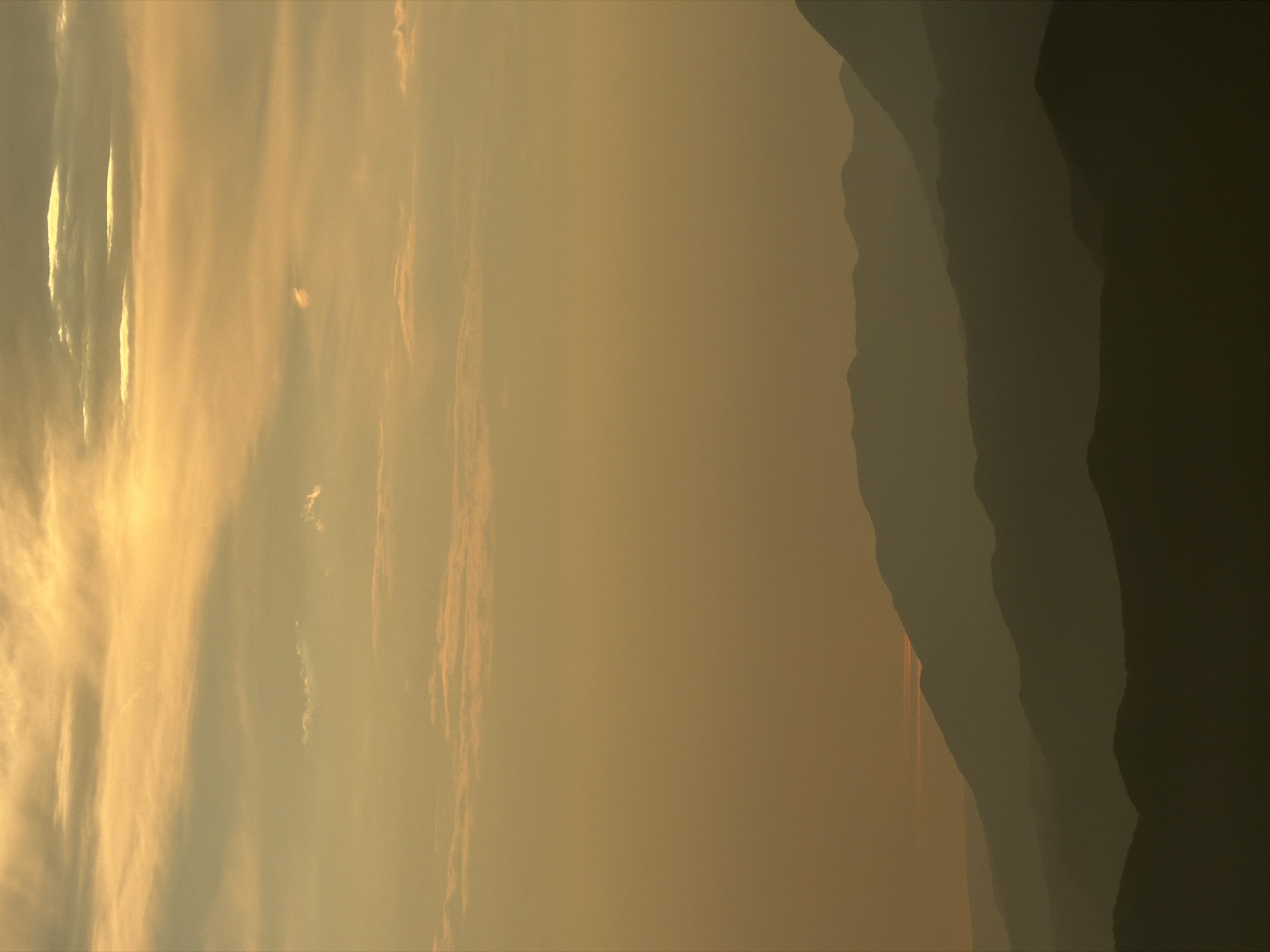 This is a photograph of a mountain range at dusk or dawn. The foreground is dominated by several layers of silhouetted mountains, creating a sense of depth and scale. The mountains are dark and indistinct, their shapes softened by the atmospheric haze. Above the mountains, the sky is a gradient of warm colors, ranging from a pale yellow at the horizon to a soft orange and then a lighter yellow at the top. Wispy clouds are scattered across the sky, illuminated by the setting or rising sun. The overall impression is one of serene natural beauty and tranquility. The image does not depict any specific location within the city of Kuk, Montenegro, as it focuses on a natural landscape. There are no people, objects, or actions visible in the scene.