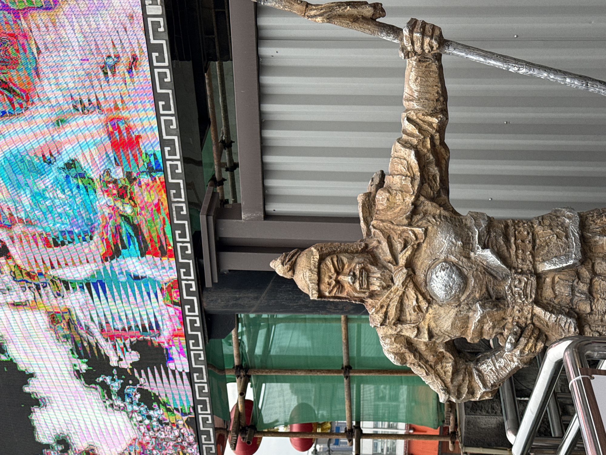 A detailed close-up shows an architectural detail featuring a sculpted, brown-colored figure hanging from a metal bar. The figure has a somewhat weathered appearance, suggesting it may be an older piece or a replica designed to look aged. The sculpture's head is prominent, with detailed facial features and a cap-like head covering. The figure is hanging upside down with its left hand wrapped around the bar.
Behind the figure, there are construction-related elements such as scaffolding covered with green netting. Parts of an advertisement are visible on the left. The background includes corrugated metal siding, typical of construction or renovation sites.
Based on the prompt, the location is Yuzhong District, China.