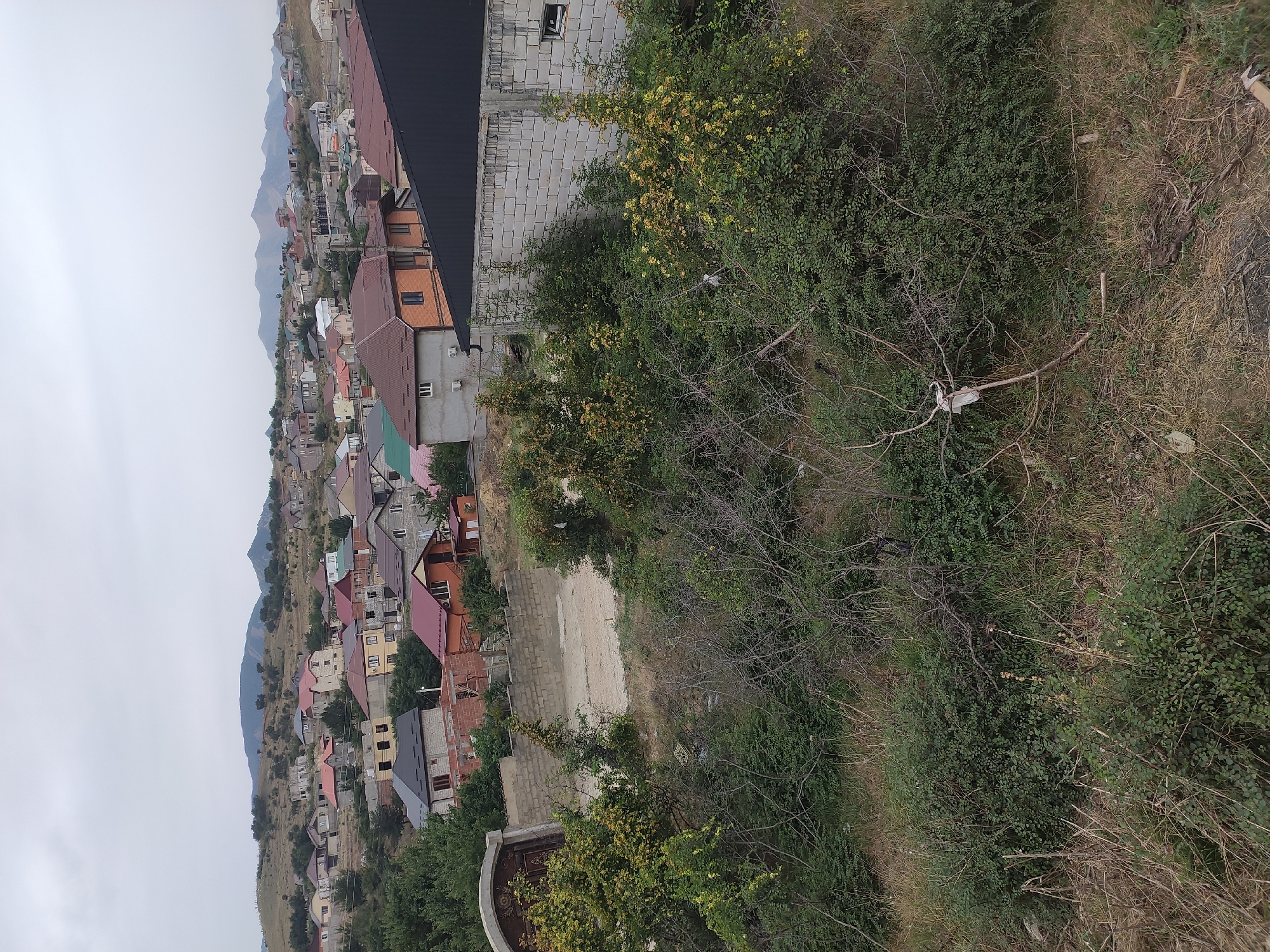 This is a high-angle, wide shot of a residential area in the city of Makhachkala, Russia. The image is overcast and features a dense collection of houses with red and brown tiled roofs, interspersed with construction sites where new homes are being built. The houses vary in architectural style, with some being multi-story with balconies and others appearing to be single-family dwellings. In the foreground, there is thick, overgrown vegetation with a scattered white object on a branch. A concrete wall with a large arched gateway is visible on the left side of the image. Beyond the houses, a chain of low, rolling hills stretches into the distance under a cloudy sky. The overall scene depicts a developing urban landscape within a natural, hilly environment.