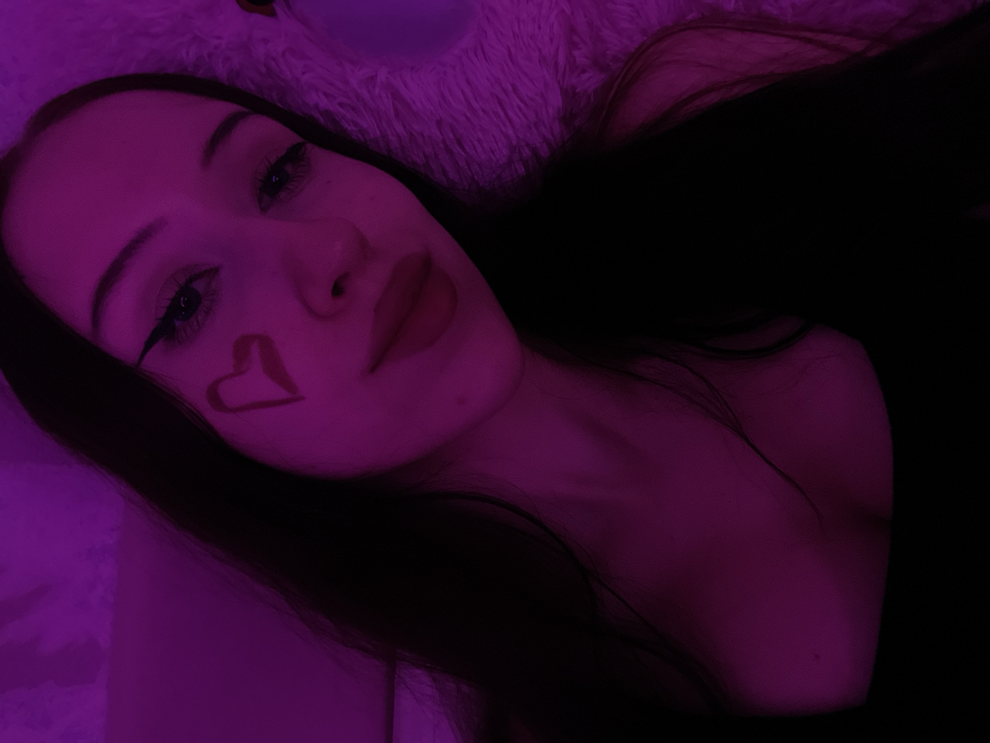 A young person with long dark hair, wearing a dark top, is pictured in a close-up, head-and-shoulders shot. They have a heart drawn on their cheek and are wearing winged eyeliner. The lighting is predominantly purple, creating a dramatic effect. The background appears to be a fluffy, textured surface, possibly bedding or a blanket. There are no discernible elements that indicate a specific location within the city of Votkinsk, Russia. The image captures a self-portrait with artistic makeup and lighting.