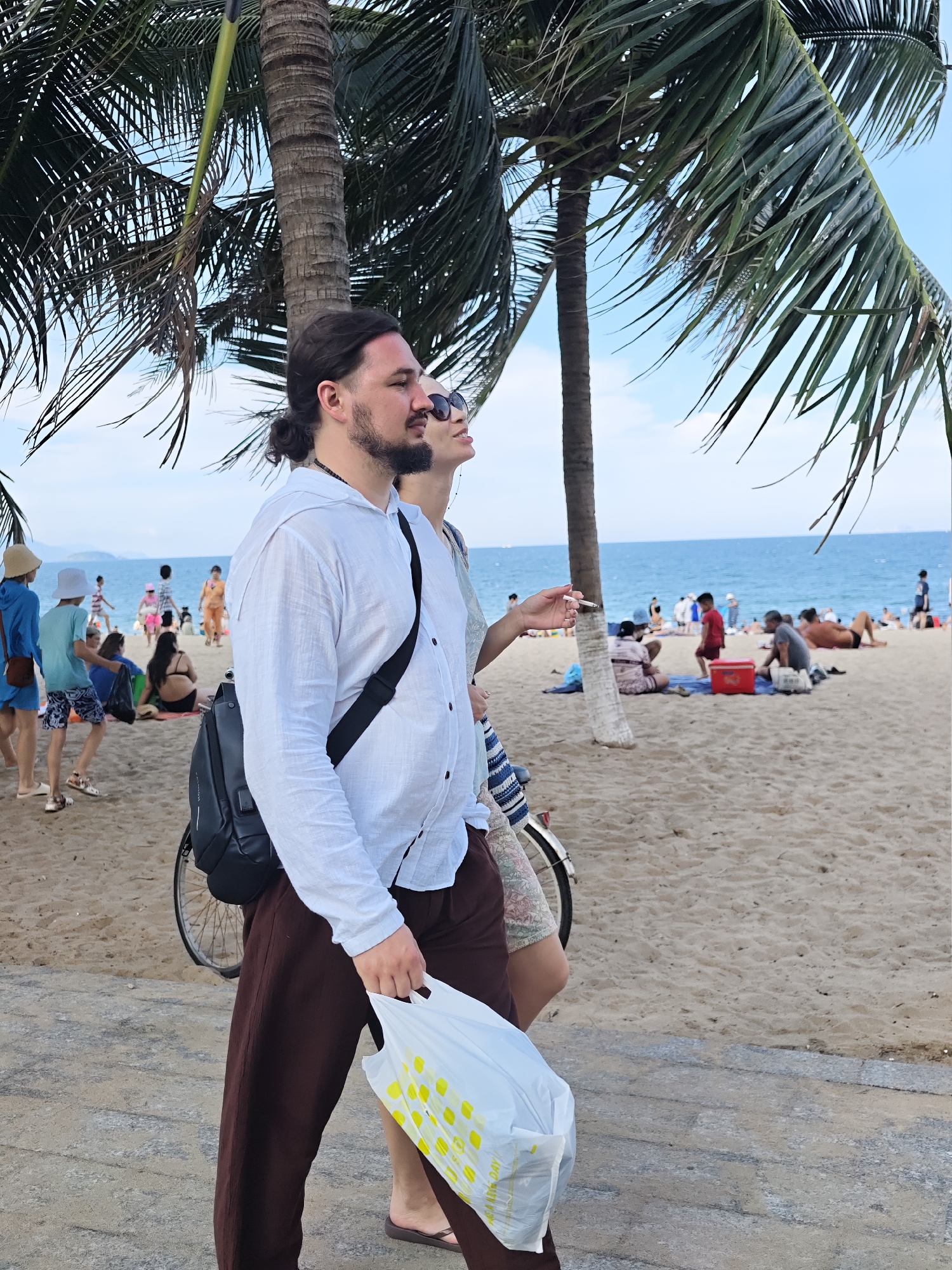 This image depicts a beach scene in Nha Trang Ward, Vietnam, on a sunny day. In the foreground, a man with dark hair tied back in a bun, a beard, and a white collared shirt walks towards the right, carrying a white plastic bag with yellow polka dots in his right hand. He has a black backpack slung over his left shoulder. Beside him and slightly behind, a woman with sunglasses and a light-colored dress looks upwards and holds a cigarette in her left hand. The background shows a sandy beach with numerous people enjoying the day. Some are sitting on towels, while others are walking or standing near the water. Palm trees with green fronds are visible in the foreground and mid-ground, casting shadows on the sand. The ocean is a vibrant blue, meeting the horizon under a clear sky. A bicycle is partially visible behind the man.