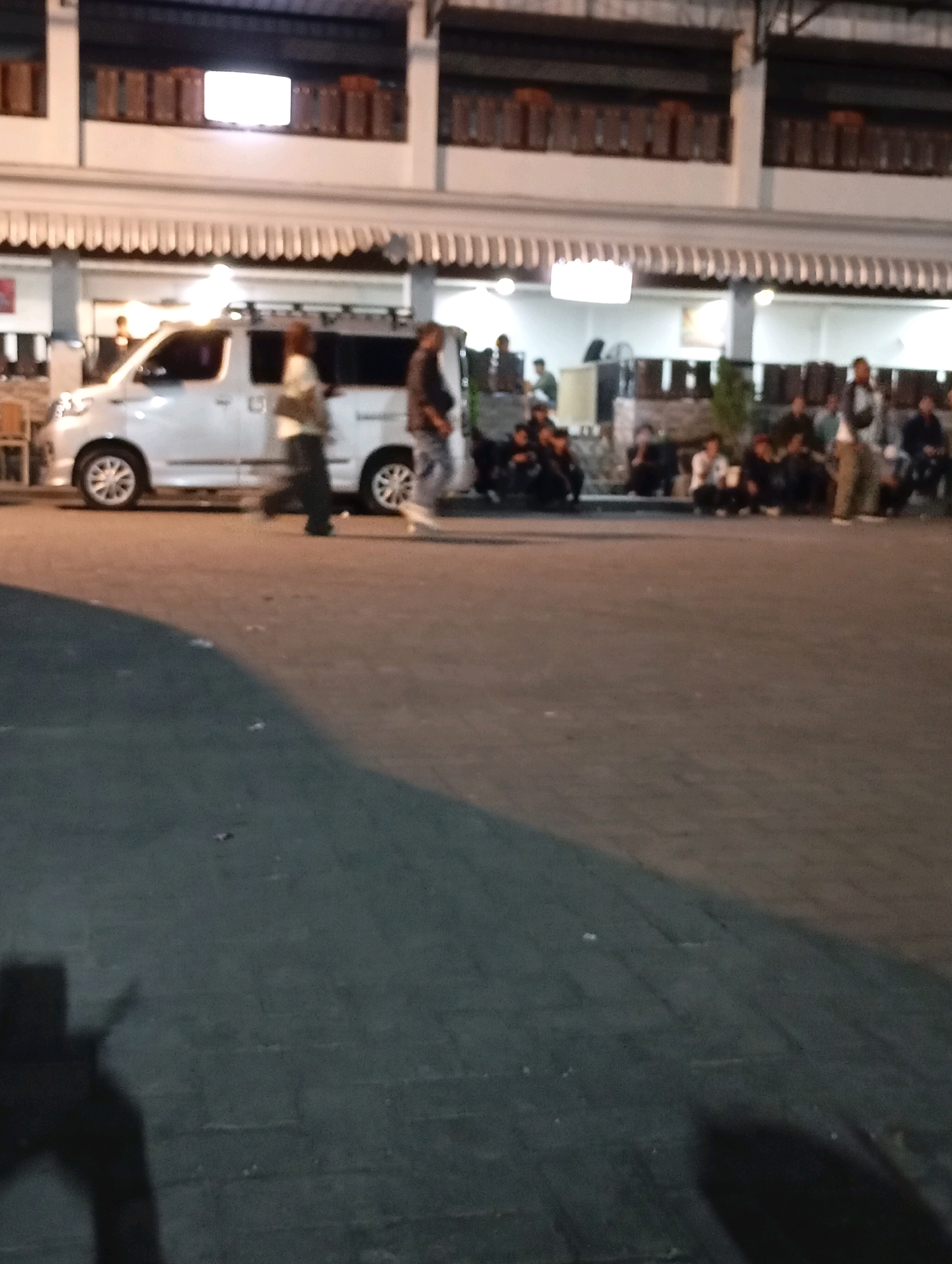 In Purworejo, Indonesia, on a paved street at night, a white van is parked. Two individuals are walking near the van. In the background, a group of people are seated. The scene appears to be in front of a building with awnings.