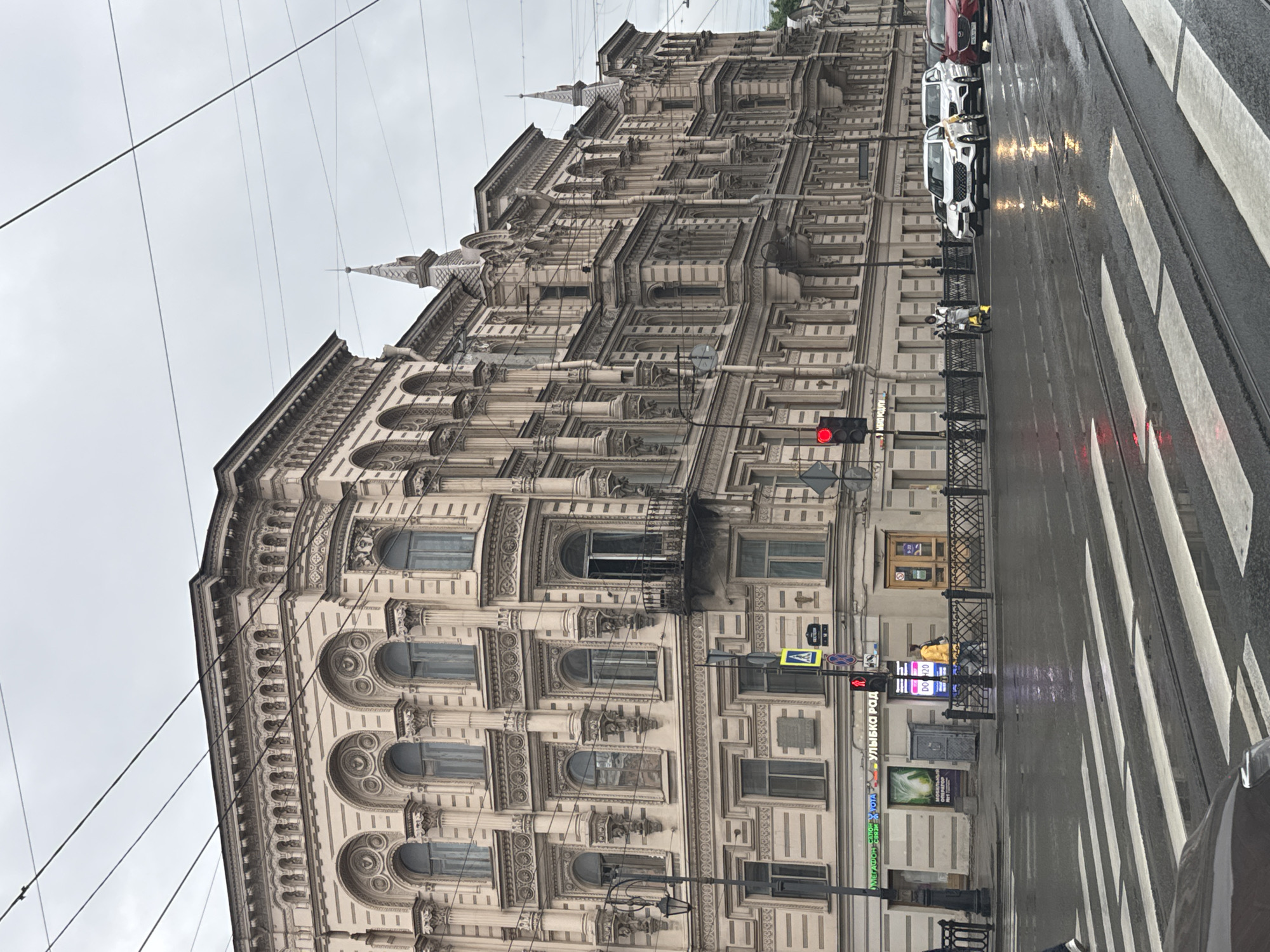 The image depicts a street scene in Saint Petersburg, Russia, on a rainy day. The architecture suggests a historic district, with ornate buildings featuring multiple stories, arched windows, balconies, and decorative facades. The street is wet and reflects the overcast sky and traffic lights.
Several cars are visible on the wet road, including white SUVs and a red sedan. A white SUV with yellow accents and a driver wearing a yellow vest is stopped at a traffic light. Another vehicle, partially visible in the foreground, is also on the road. Pedestrians are present, including a person in a yellow jacket walking across the street, and another individual on a yellow scooter or bicycle.
A traffic light shows a red signal, indicating a stop. Street signs and a pedestrian crossing are also visible. The buildings house various businesses, indicated by signage such as "МЕГАФОН" and "УЛЫБКА РАДУГИ". Overhead wires, likely for public transportation or utilities, crisscross the sky. The overall atmosphere is somber due to the cloudy weather and wet conditions.