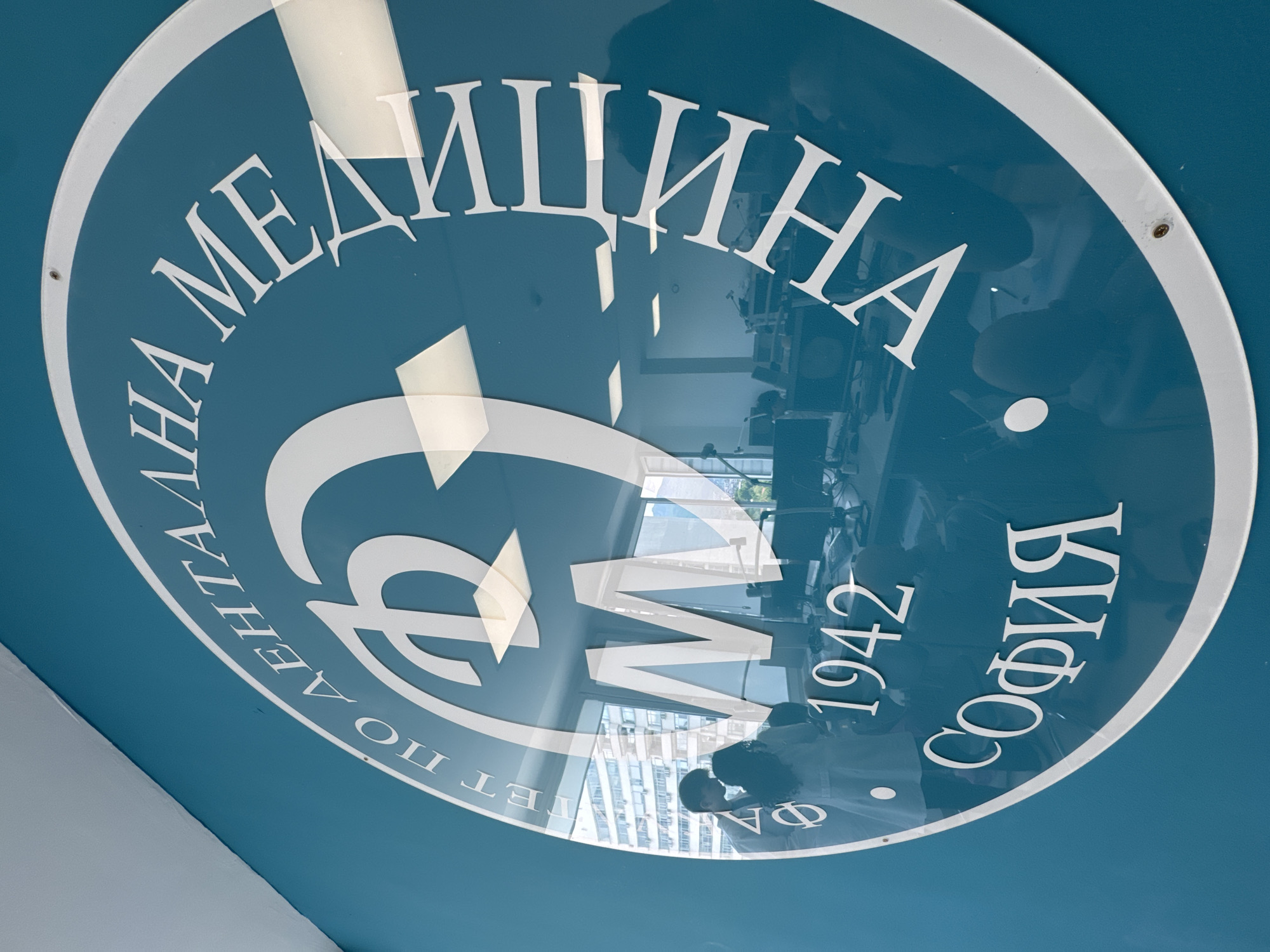 The image shows a circular sign in the city of Sofia, Bulgaria, belonging to the Faculty of Medicine. The sign is predominantly blue with white lettering in Cyrillic. The words "Медицина София" are visible along the upper and lower arcs of the circle, respectively, with "1942" indicating the founding year. A stylized symbol, possibly a caduceus or similar medical emblem, is positioned in the center of the circle. Reflections in the sign indicate a contemporary indoor environment with people present.