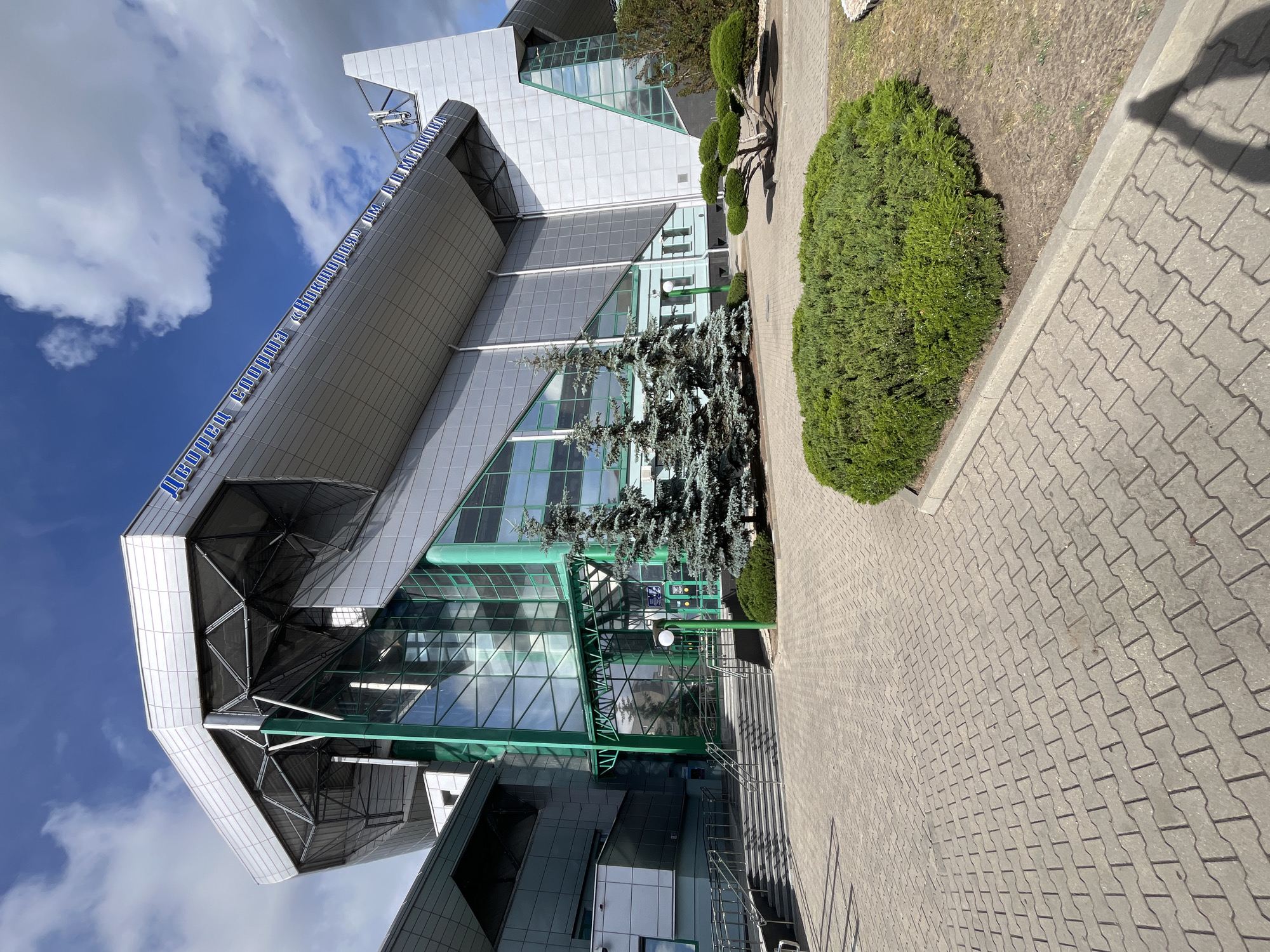 The image shows a large, modern building with a silver, angular facade and green-framed glass entrances. The building has a sign in blue lettering that reads "Дворец спорта «Виктория» им. А.М.Ломакина". In front of the building, there is a paved walkway made of interlocking gray pavers. Several evergreen trees, including two tall, slender blue spruces and a small, sculpted evergreen tree, are planted in the foreground. A large, dense green bush is also visible. The sky above is blue with scattered white clouds, and the sun is casting shadows on the pavement. There are no people visible in the image. The scene appears to be an exterior view of a sports arena or complex.