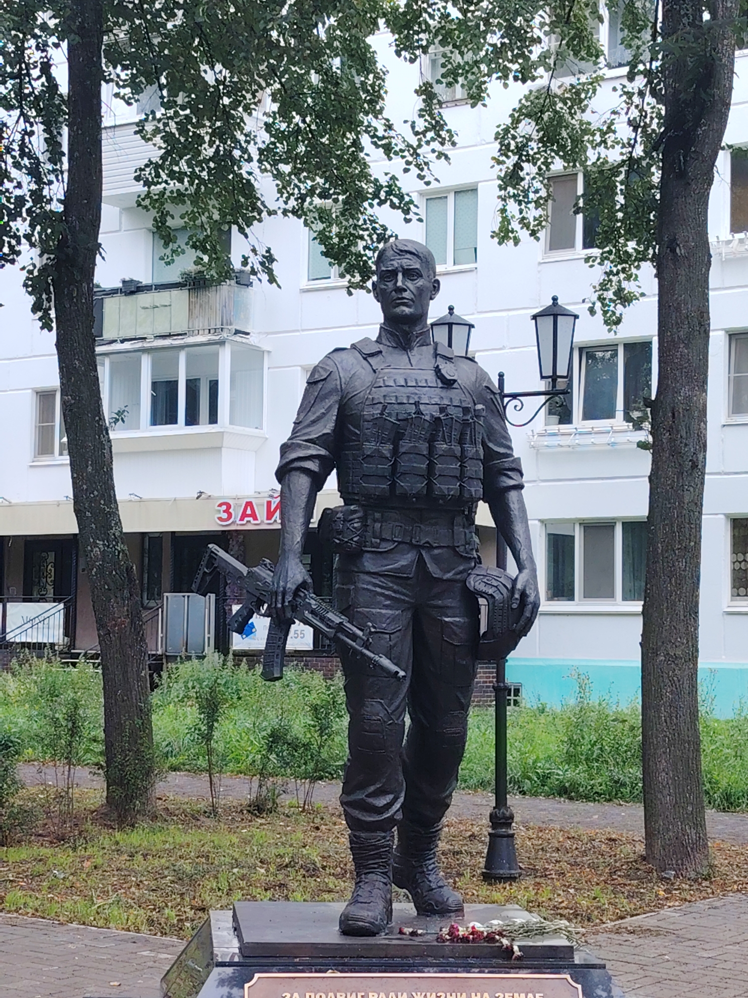 A statue of a soldier stands in front of a white building in Chaikovsky, Russia. The soldier is depicted wearing tactical gear, including a bulletproof vest, and is holding a rifle in his right hand and a helmet in his left. The building behind the statue appears to be an apartment complex with balconies and windows. Trees with green leaves frame the statue, and a lamppost is visible to the right of the soldier. A sign with Russian lettering, "ЗАЙ", is visible on the building. The base of the statue has an inscription in Russian. The ground in front of the statue is paved with bricks, and there is a patch of grass and bushes. Flowers are placed at the base of the statue.