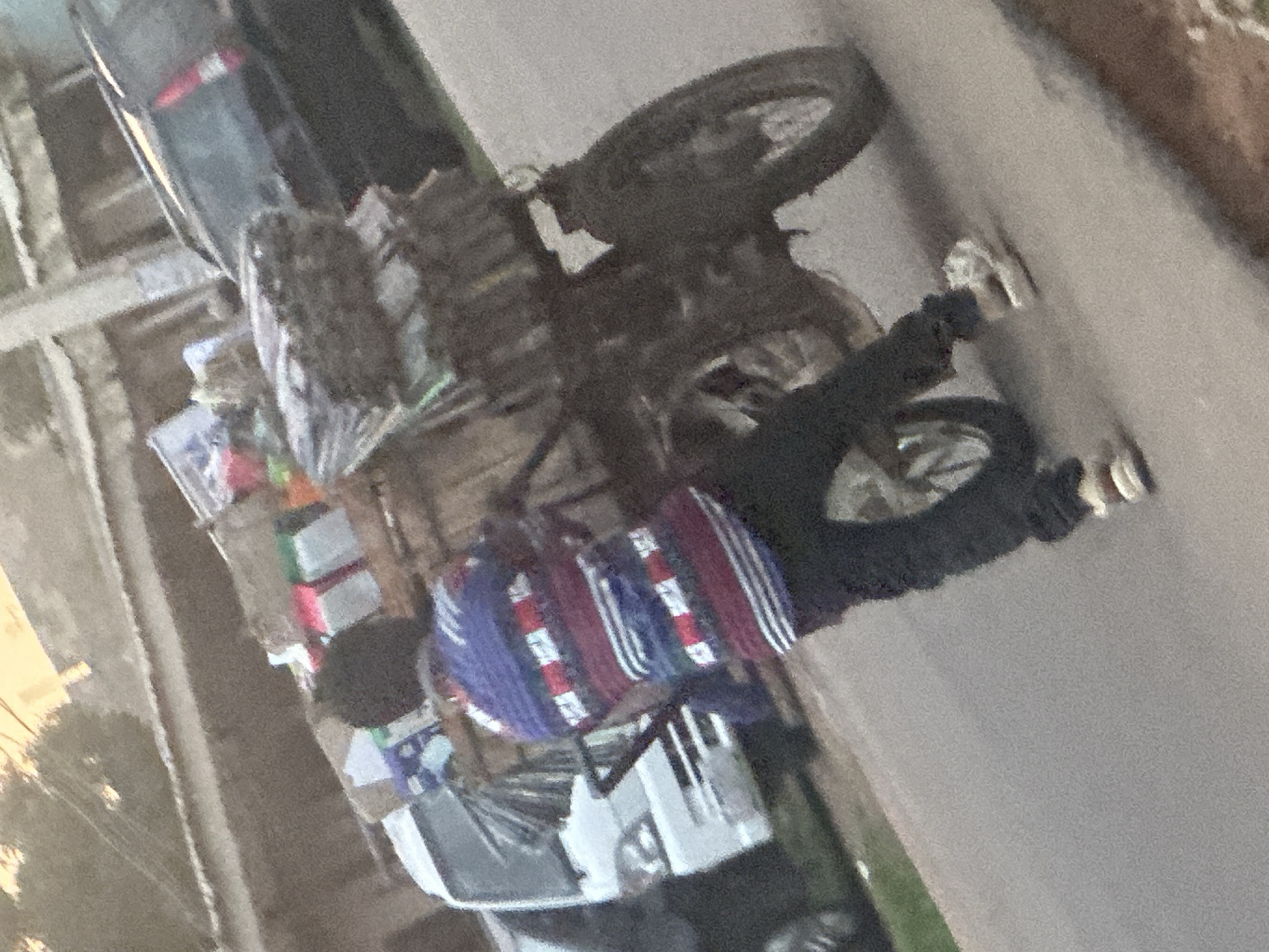 In Jos, Nigeria, a person wearing a striped blue, red, and white shirt and black pants is standing next to a motorcycle laden with goods. The motorcycle is facing to the right of the frame, and the person is facing away from the camera, looking towards the motorcycle. The goods on the motorcycle are stacked high and appear to be various items, possibly packaged goods and bundles of material. A white commercial vehicle is parked to the left of the motorcycle. The scene is on an asphalt road, with a blurred background of buildings and overhead wires, suggesting an urban or semi-urban environment.