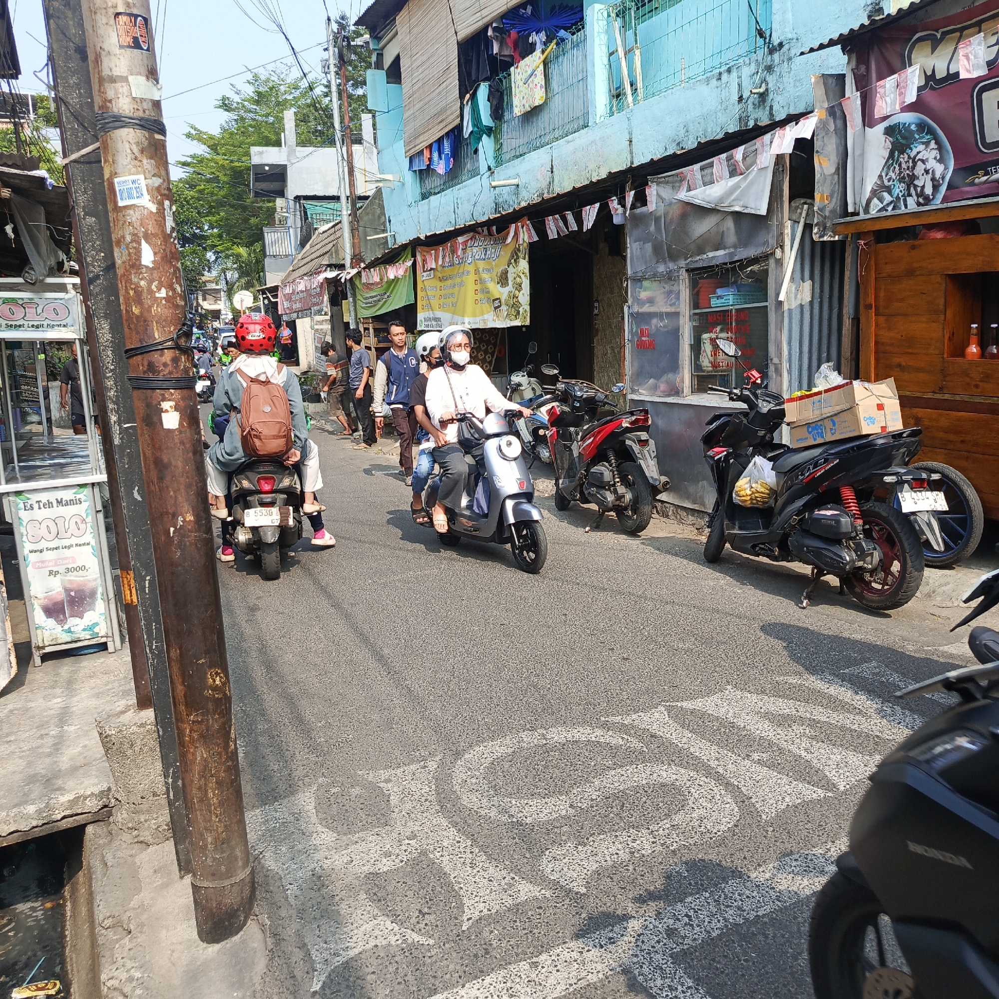 The image depicts a street scene in South Jakarta, Indonesia, likely in a densely populated urban area. In the foreground, a utility pole stands prominently on the left side of the frame, its surface adorned with various stickers. The street is paved with asphalt and shows signs of wear.
Several individuals are present, primarily riding motorcycles. Two people are on a silver scooter in the center of the image, with the person in the front wearing a white shirt and a face mask, and the person behind wearing a red helmet and a brown backpack. Another motorcycle with a red frame and black accents is parked on the right, with boxes stacked on its rear seat. A black motorcycle, partially visible in the lower right corner, also appears to be parked.
The surrounding environment features buildings with blue and light blue facades, some with corrugated metal roofing. Clothes are seen drying on a balcony of one of the buildings. Various signs and banners are displayed on the storefronts, including advertisements for food items like "Nasi Goreng" and "Es Teh Manis SOLO." The scene suggests a commercial and residential area with a mix of activities. The presence of Indonesian license plates and written Indonesian text on signs confirms the location. The overall impression is of a bustling, everyday street in an Indonesian city.