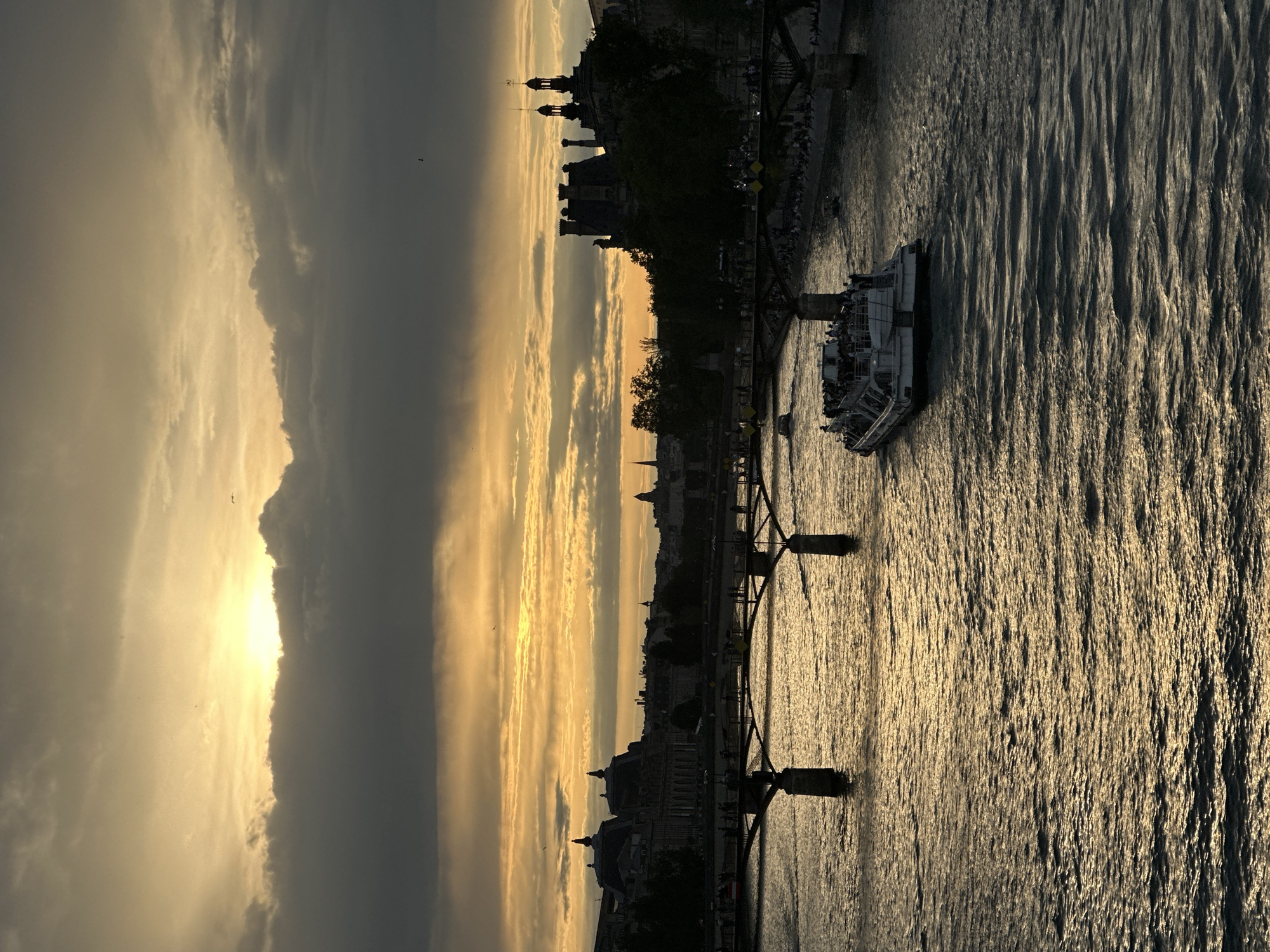 This photograph captures a sunset view over the Seine River in Paris, France. A tour boat with people on board is on the water. A portion of a bridge is visible, with the silhouettes of Parisian buildings including the Musée d'Orsay visible along the riverbank. The sky is filled with clouds and sunlight.