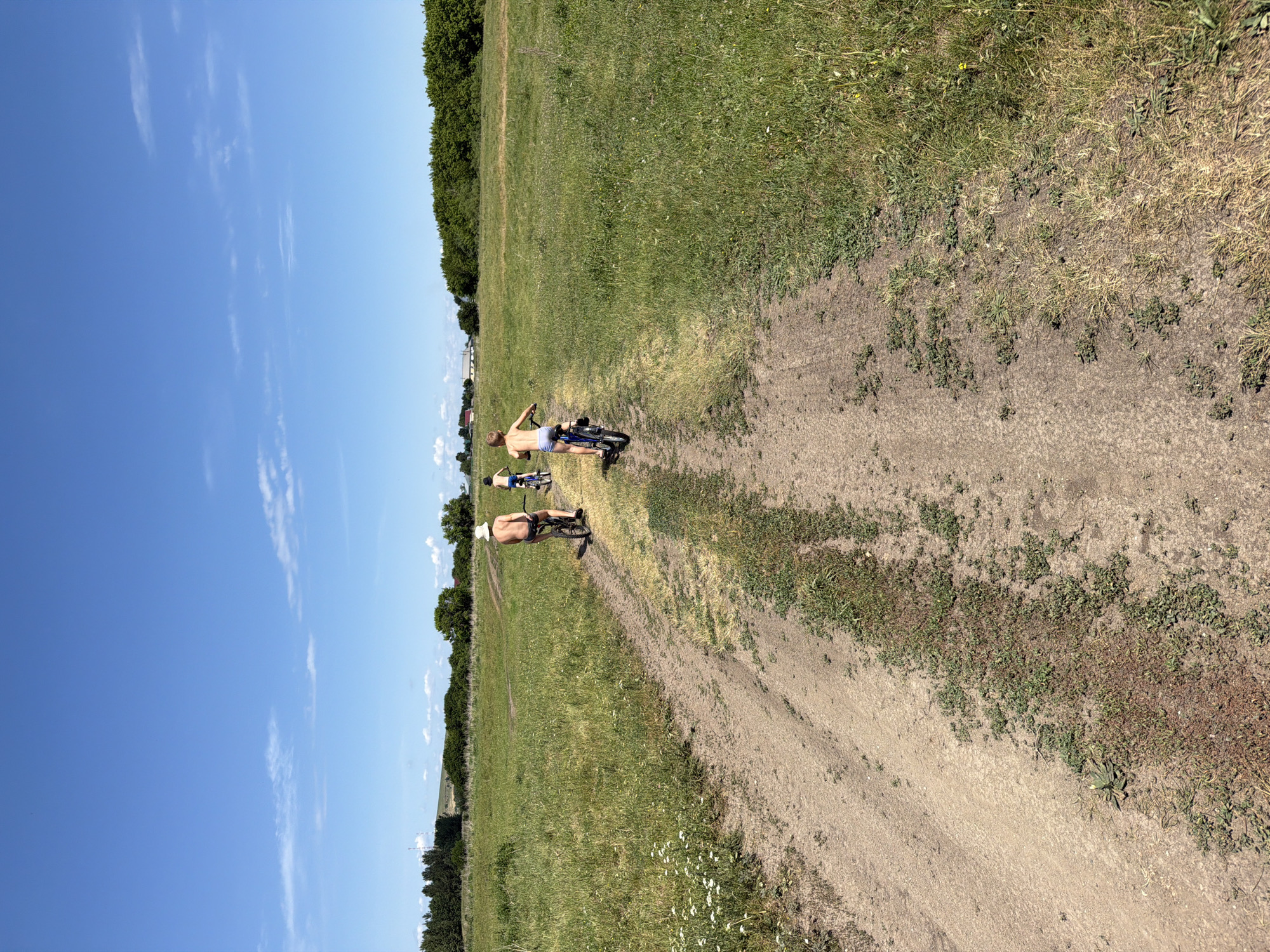 The image depicts a sunny day in Zelenyi Dol, Russia, with a clear blue sky and scattered white clouds. The scene is dominated by a vast grassy field, with a dirt path winding through it. A line of dense green trees marks the horizon.
In the middle ground, two individuals are riding bicycles on the dirt path. The person in front is wearing a white hat and dark shorts, while the person behind them is wearing blue shorts. Both appear to be riding small, blue bicycles. The overall mood seems relaxed and casual, suggesting a recreational activity on a pleasant day. The sunlight indicates it is daytime, likely late morning or early afternoon. The green foliage and clear sky suggest a warm, summer day. There is no visible text in the image.