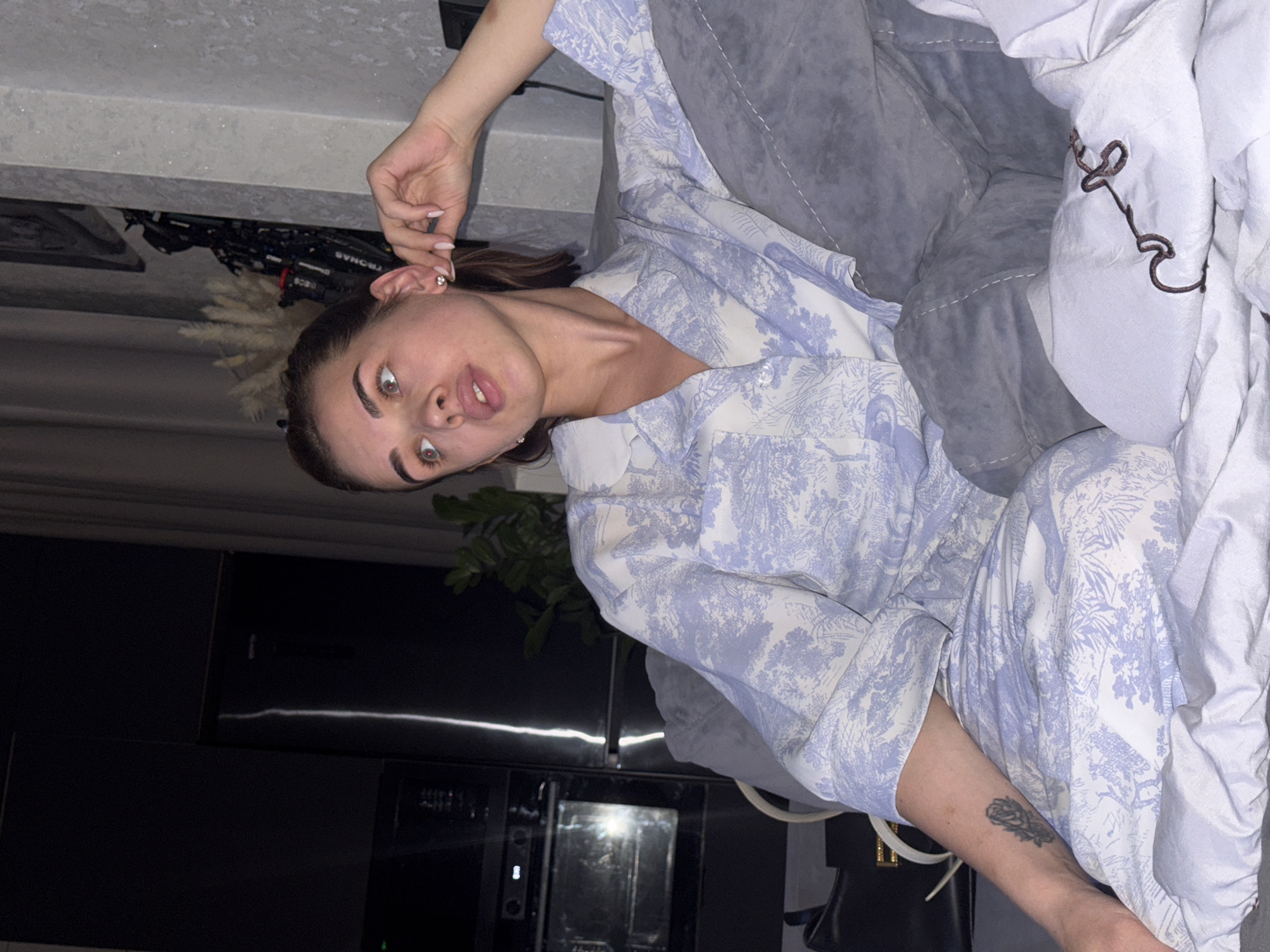 A woman is lying on a gray couch in what appears to be a modern apartment. She is wearing blue and white pajamas, and she has a tattoo on her left forearm. Her face is toward the camera and she is sticking her tongue out. The apartment includes a black kitchenette area visible in the background. A houseplant stands next to the kitchenette. The location is Лобаново, Russia.