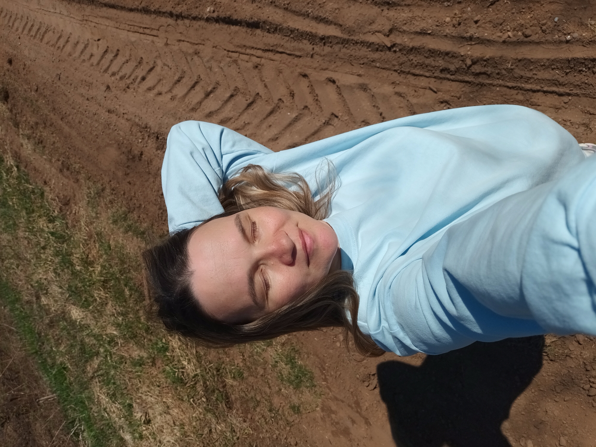 The image shows a woman lying on the ground. She has fair skin and light brown hair with some blonde highlights. Her eyes are closed and she appears to be relaxed. She is wearing a light blue long-sleeved shirt. The ground she is lying on consists of dry, brown soil with some patches of green grass. There are tire tracks visible in the soil. A shadow is cast in the lower right corner of the image. The location is identified as Ольховка, Russia.
