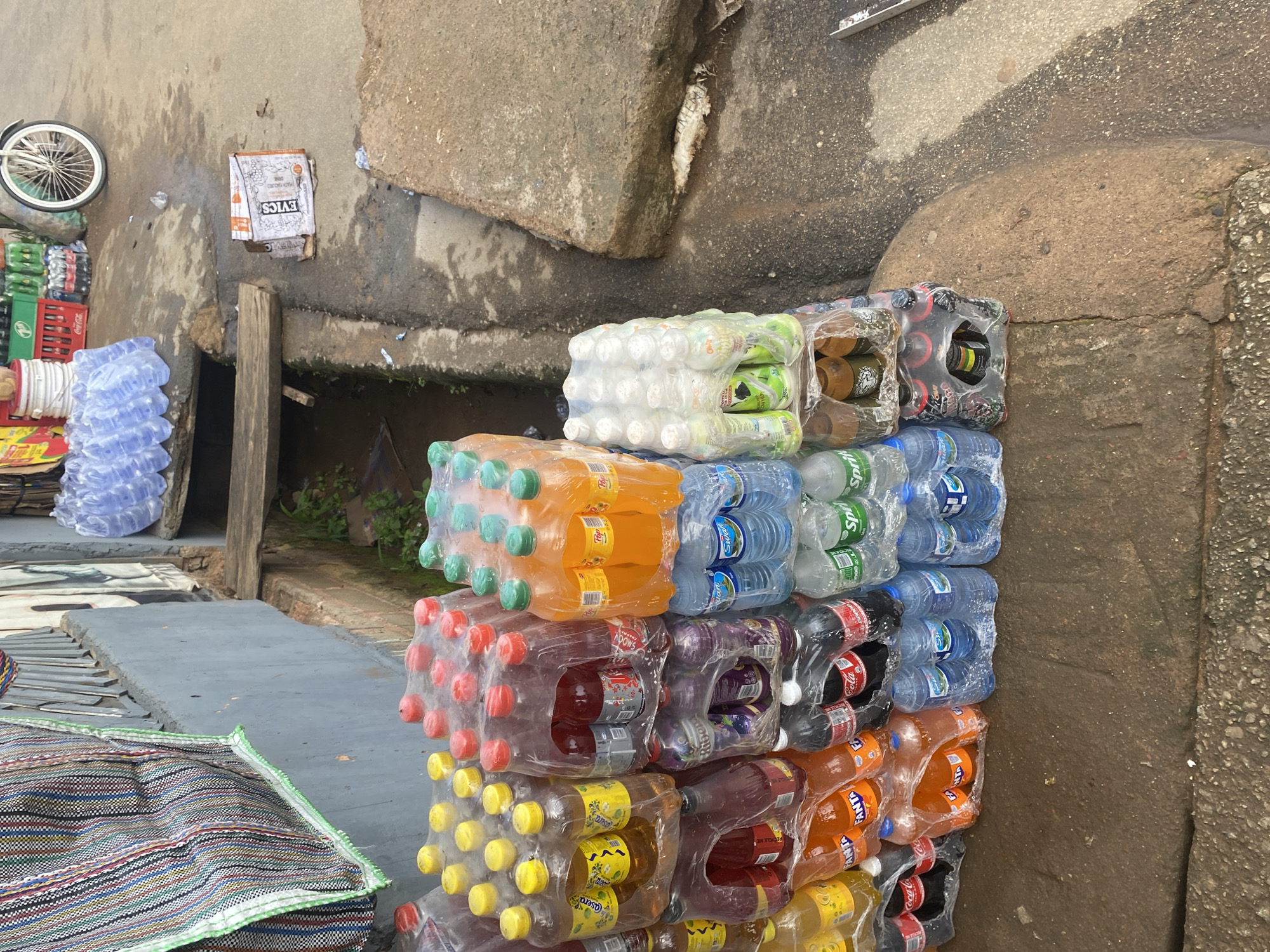 The image displays a street scene in Jos, Nigeria, where various beverages are neatly stacked for sale. The primary subjects are stacks of bottled drinks, predominantly soft drinks and water, packaged in clear plastic. These are arranged in multiple rows and columns, showcasing a variety of colors from the liquids within – green, orange, red, yellow, and brown. Some bottles are clear, containing water.
The setting appears to be an outdoor market or roadside stall. In the background, there are rough, concrete structures, possibly walls or large stones, and a wooden support beam. To the left, a bicycle wheel is visible, suggesting a mode of transport or perhaps part of the vendor's setup. There's also a glimpse of colorful woven bags on the ground. A white and orange cardboard box with visible text is also present.
The activity is the display and likely sale of these beverages. The scene suggests a daytime setting, likely during market hours. There are no people actively depicted in the frame, but the setup implies a commercial transaction is taking place or about to. The lighting suggests it's not the harsh midday sun, perhaps morning or late afternoon.
Notable details include the clear shrink-wrapping of the drink packs, keeping them organized and clean. Text is visible on some of the packaging. For instance, the orange-colored drinks clearly show the "FANTA" logo. Other labels might be visible upon closer inspection of the different drink packs. The overall impression is one of a typical, bustling informal market environment.