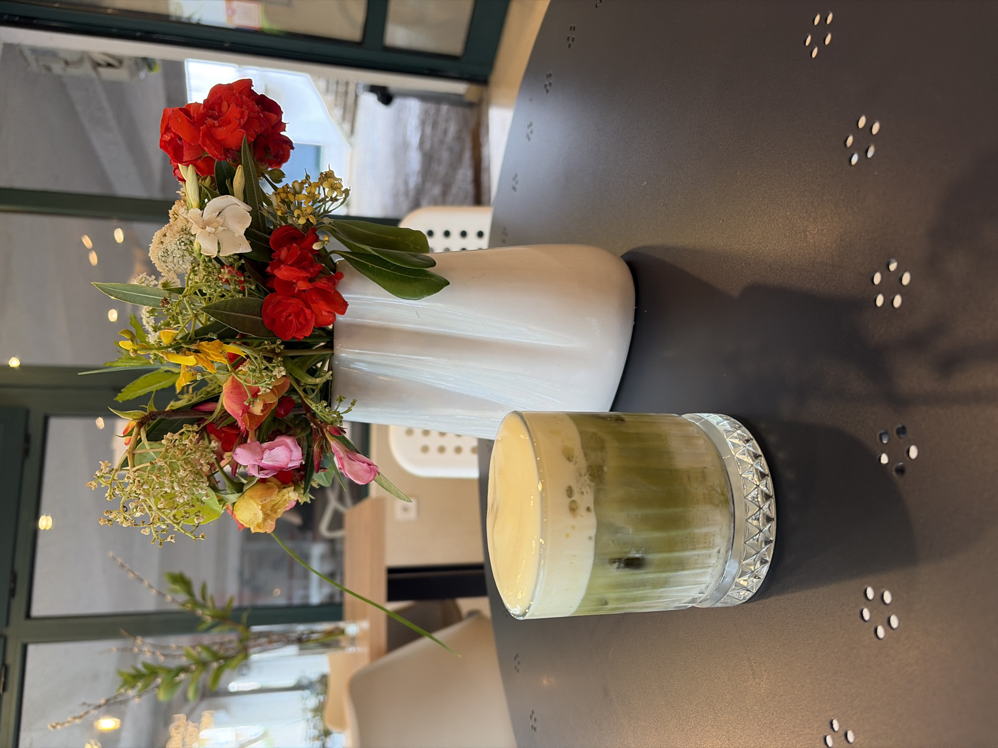 The image shows a table arrangement with a vase of flowers and a glass of a green drink, possibly matcha latte. The flowers are a mix of red, white, yellow, and pink blooms with greenery, arranged in a white, ribbed vase. Next to the vase is a short glass with what appears to be a creamy, foamy top layer over a green beverage. The table is dark gray with small, inlaid circular patterns. The backdrop is a bright interior, with a window and a partial view of chairs. Without additional information, I cannot confirm the location in Budva, Montenegro, or provide details about people or interactions within the scene.