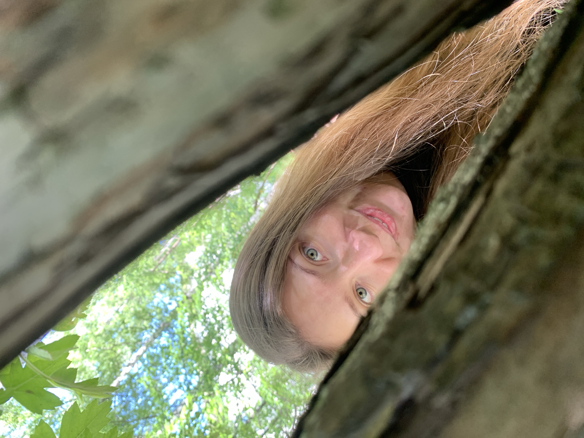 A woman with light skin, blue-green eyes, and light brown hair is looking down through a narrow gap between two rough, gray-brown wooden surfaces. The background seen through the gap shows green leaves and glimpses of a bright, somewhat blurry sky, suggesting trees overhead. The woman's face fills most of the visible space within the frame. Location details (Chaikovsky, Russia) are not discernible from the image.