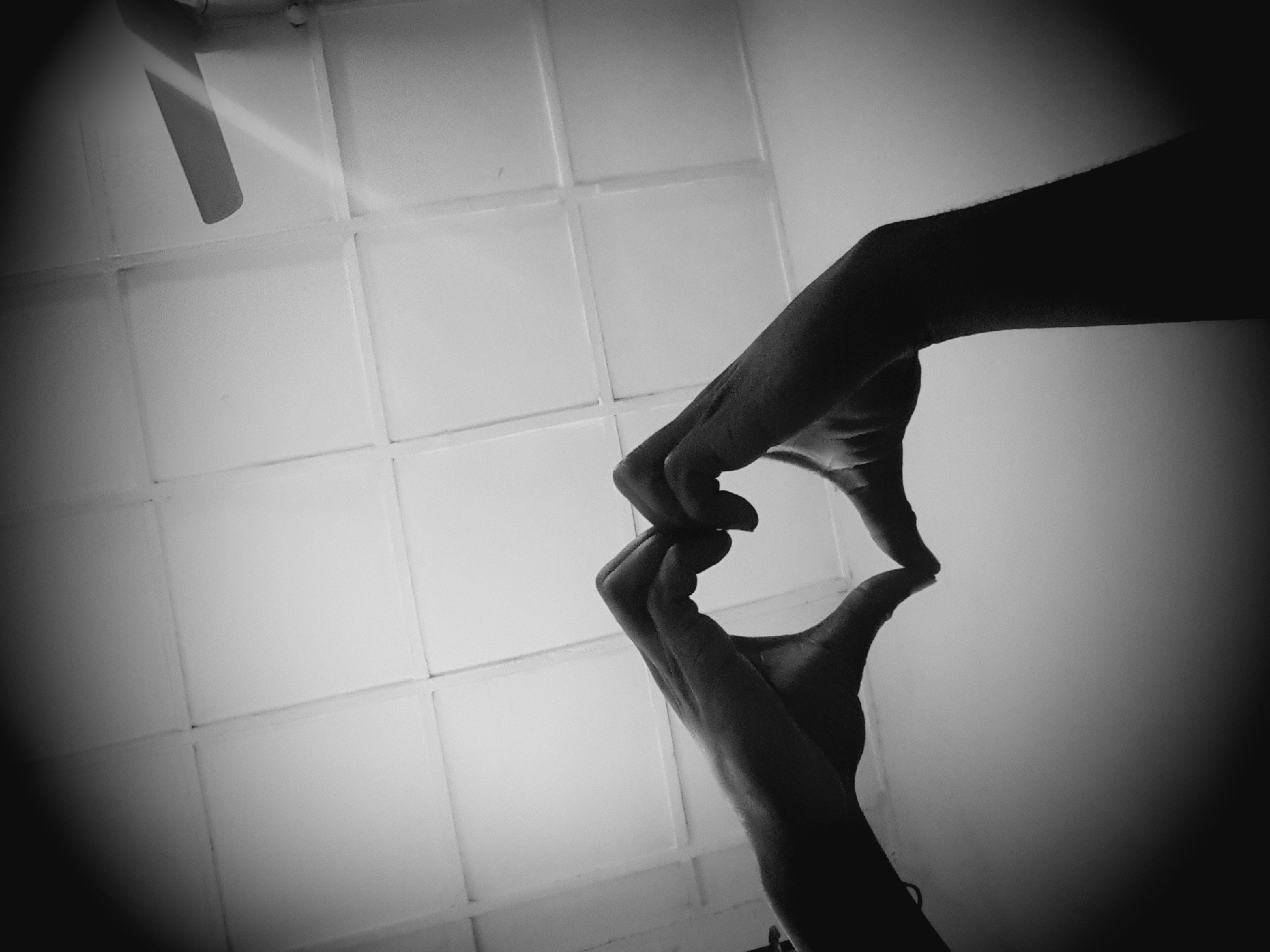 A monochromatic photograph, captured from a low-angle perspective looking upward, depicts two human hands silhouetted against a bright background, forming a heart gesture. The hands are positioned centrally in the lower half of the frame, with fingers and thumbs interlocked to create the characteristic heart shape. The background consists of a light-colored ceiling featuring a prominent grid pattern, suggesting square or rectangular panels or exposed beams. A portion of a light-colored ceiling fan, including one blade and part of its housing, is visible in the upper right quadrant of the frame. The image exhibits strong vignetting, with darker areas around the periphery. This scene is located in the city of Narayi, Nigeria.
