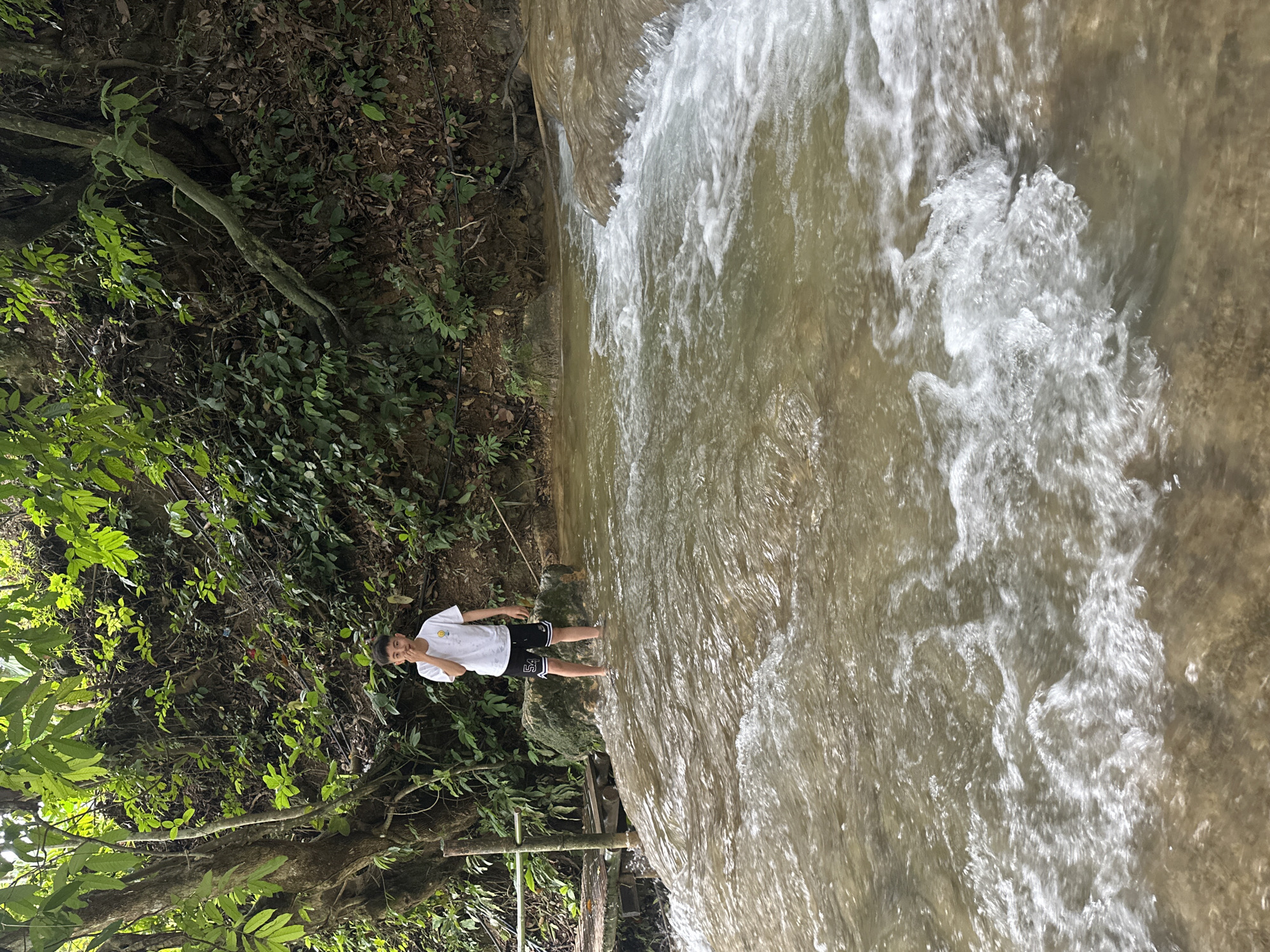 A young boy stands in a shallow, flowing body of water at a waterfall in Cổ Lũng Commune, Vietnam. The boy, wearing a white t-shirt and black shorts with white trim, is looking at the camera with his right hand raised to his face. The water is a muddy brown color and appears to be cascading over rocks. Lush green vegetation covers the hillside behind the boy, with trees and vines growing densely. A wooden structure, possibly a bridge or platform, is visible to the left of the boy, partially submerged in the water. The overall scene depicts a natural, outdoor environment.