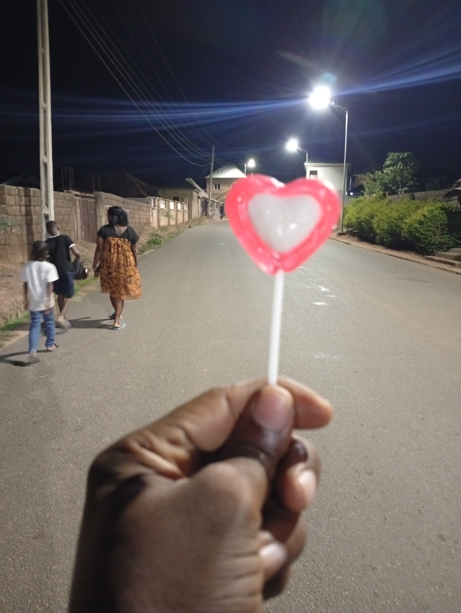The image is taken from a first-person perspective, with a hand holding a heart-shaped lollipop in the foreground. The lollipop is pink and white, with a white stick. In the background, a street stretches away under the glow of streetlights. A family consisting of a woman, a man, and a child are walking away from the viewer along the left side of the street. Houses and fences line the street, and foliage is visible along the right side. The location is Barakin Akawo, Nigeria, as determined by the context of the prompt. The time of day appears to be night.