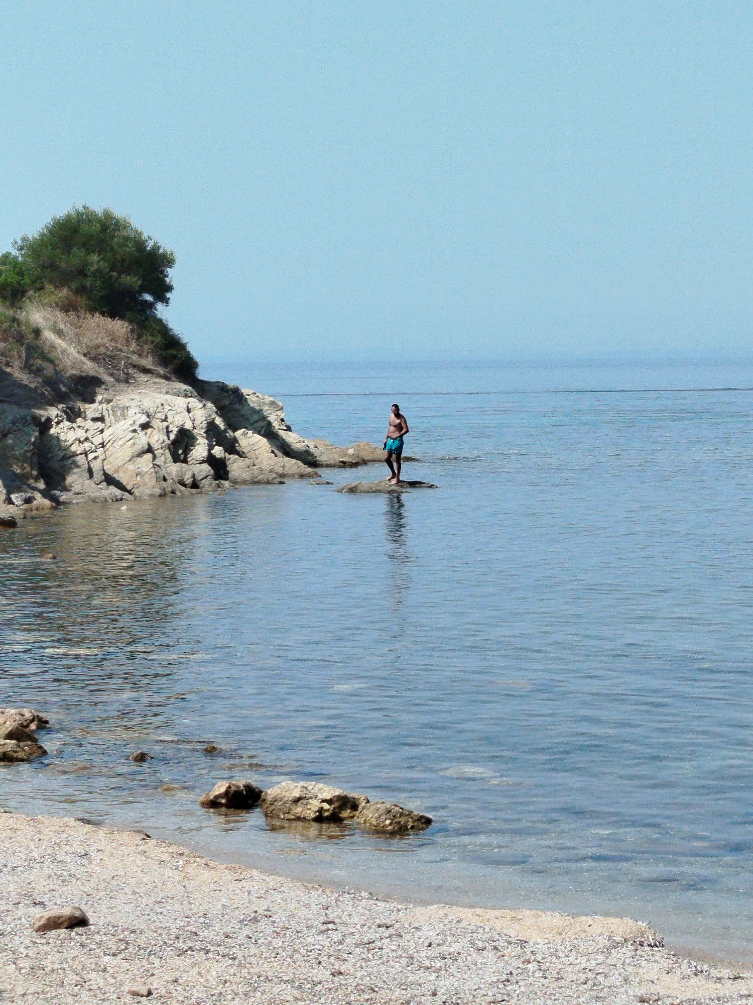 A man stands on a rock in the clear, calm sea near the coast of Sithonia Municipal Unit, Greece. The man is wearing blue swim trunks and is facing away from the camera. The sea is a bright blue, with gentle ripples. The coastline is rocky, with some vegetation growing on the higher ground. The sky is a clear, light blue. The beach is made of small pebbles.