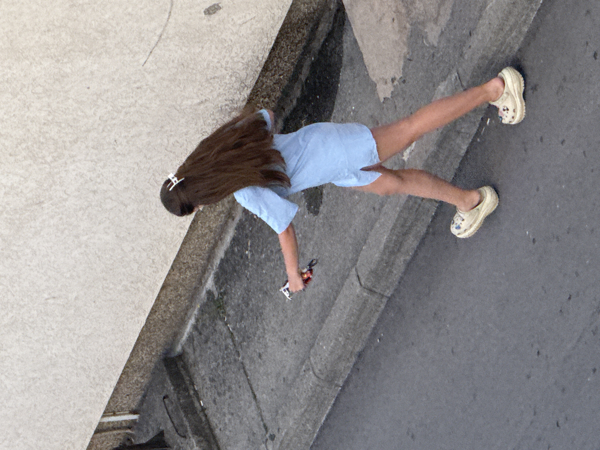 A person with long, dark brown hair, held back by a white clip, walks along a sidewalk. They are wearing a light blue short-sleeved dress and light-colored clogs with a patterned design. The person is holding a small object, possibly a vape or electronic cigarette, in their right hand. The environment appears to be a street scene in Budva, Montenegro, with a textured beige wall in the background, a concrete sidewalk, and asphalt road.
