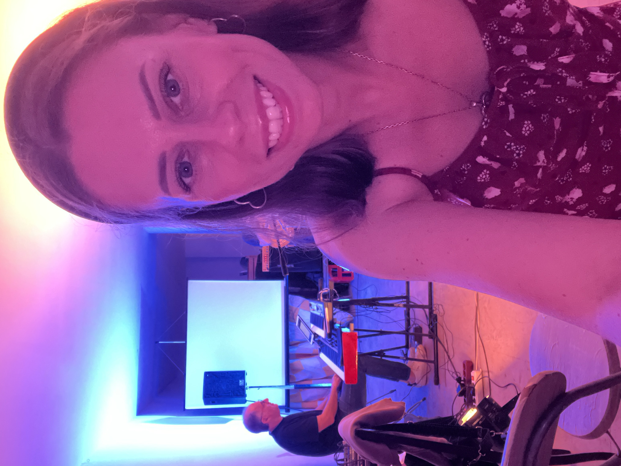 The scene depicts an indoor performance or rehearsal space in Santeramo in Colle, Italy, illuminated by dynamic colored lighting.
In the foreground, a woman is captured in a selfie-style shot, smiling directly at the viewer. She is an adult with long, dark brown hair, parted in the middle. Her facial features include defined eyebrows, eyes gazing forward, and a wide smile revealing her upper teeth. She wears a dark-colored (likely maroon or deep red) sleeveless top adorned with a light-colored, possibly white or pink, floral or abstract pattern. A delicate chain necklace with a small pendant rests on her décolletage, and she has small, heart-shaped hoop earrings. The dominant lighting on her is magenta-pink, with hints of orange and blue on her right side.
In the mid-ground, to the left of the woman, an adult man is seated on a stool, playing an electronic keyboard. He is viewed in profile, facing right. He has short, receding hair and wears dark-rimmed glasses, a dark short-sleeved t-shirt, dark pants, and light-colored sneakers. His hands are positioned over the keys of a multi-tiered keyboard, which features a prominent red lower section. Above the keyboard, a stand holds at least two electronic tablets, presumably for sheet music or control. To his left, a black monitor speaker rests on a black tripod stand.
The background features a large white projection screen or backdrop, supported by a stand. Behind this screen, a patterned fabric or curtain in beige and white is partially visible. Further to the right in the background, obscured by the foreground subject and the man's setup, additional audio equipment, possibly amplifiers or mixers, are discernible.
The environment is characterized by distinct artificial lighting. The upper left corner and ceiling areas are bathed in vibrant blue and purple light, which transitions to pink and magenta towards the right side of the frame. The floor is a light-colored, possibly concrete, surface, reflecting the varied light hues, with some orange-red light visible in the lower center. Numerous black audio and power cables are visible on the floor, connecting various pieces of equipment, including a white and red power strip. A small stage light or lamp is visible in the lower-left corner, positioned near a partially visible wooden chair with a light-colored fabric draped over it.