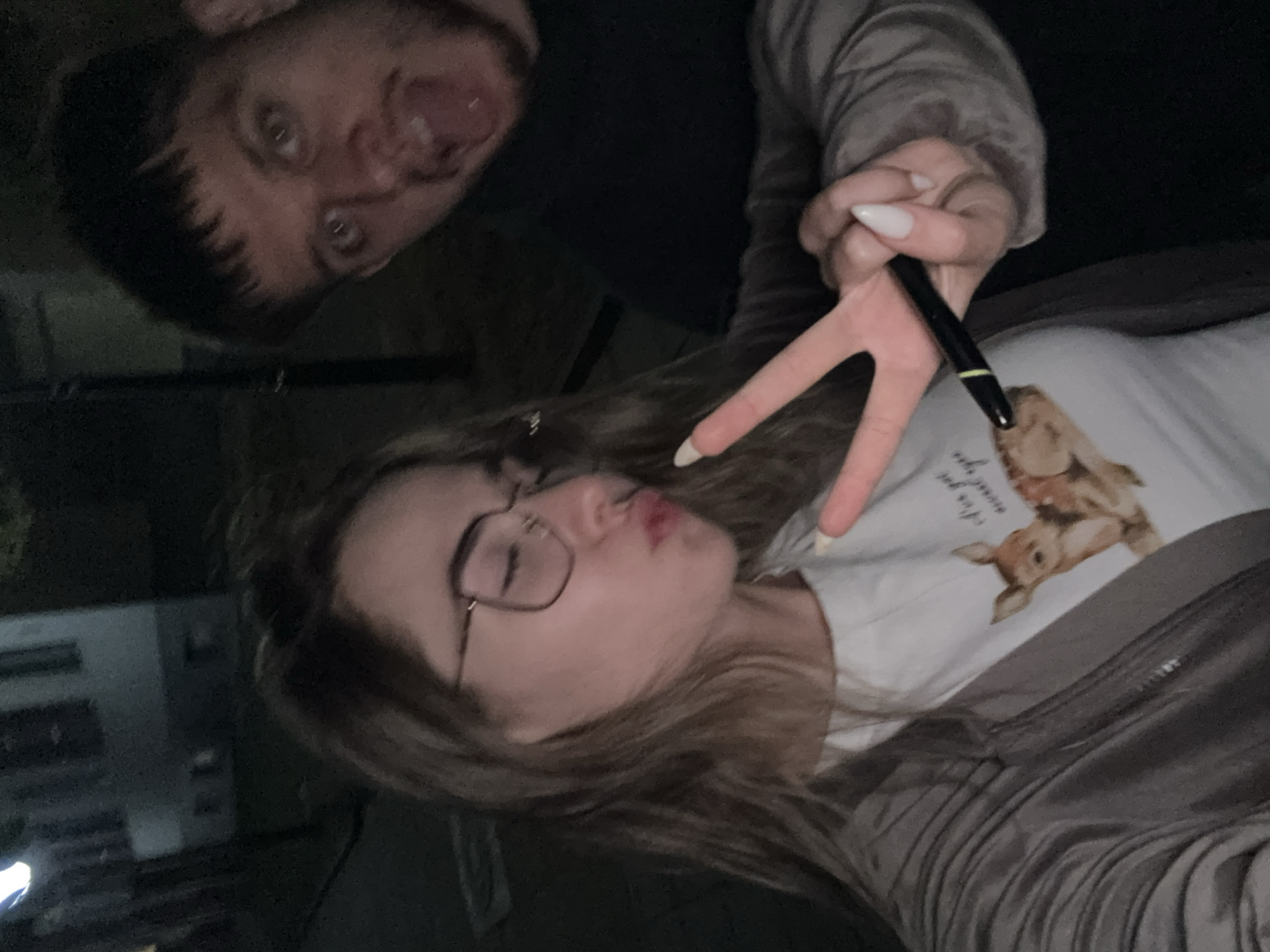 Two individuals are present in the image. The first individual, likely male, has dark hair and is positioned at the top of the frame, making a face by sticking out his tongue. The second individual, appearing female, is in the lower portion of the frame. She has light-colored hair, wears glasses, and is puckering her lips. She is holding up a pen and making a peace sign with her fingers. She is wearing a light-colored shirt with a graphic featuring what looks like rabbits. The environment is dark, and the background is nondescript, although there appears to be the vague outline of a building at the left. The location of Blagoevgrad cannot be determined from the image.
