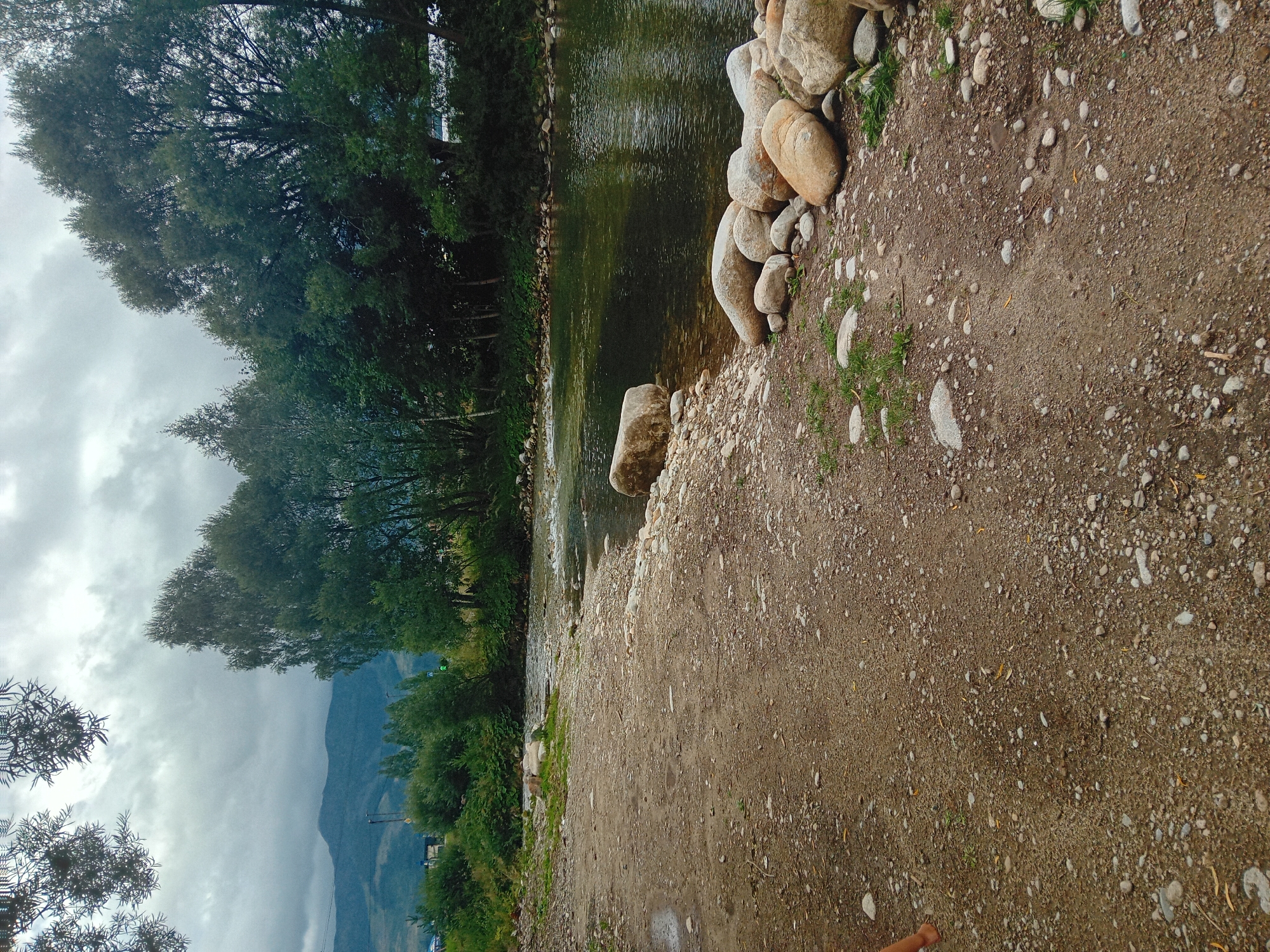 The image depicts a natural scene of a riverbank in Habovka, Slovakia. In the foreground, a dirt and gravel path leads towards the water. Numerous stones, ranging in size from pebbles to larger rocks, are scattered along the path and the river's edge. The river itself appears clear with a slight current, and its banks are lined with lush green vegetation and trees. In the background, a dense forest of deciduous trees rises, with a backdrop of rolling hills or mountains under a cloudy sky. The overall impression is one of a serene, undeveloped natural environment.