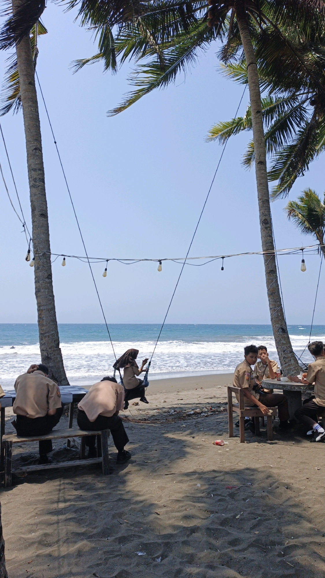 A group of young people, likely students in uniform, are gathered on a sandy beach in Desa Batukaras, Indonesia, near the ocean. Two individuals are seated on a wooden bench, facing away from the camera, under the shade of palm trees. A third person, wearing a hijab and a uniform, is sitting on a swing suspended from wires strung between two palm trees. Further to the right, a table with several chairs is set up, and three other individuals in uniform are seated around it, engaged in conversation. The ocean is visible in the background with waves breaking on the shore. String lights are hung between the palm trees, suggesting a casual outdoor dining or relaxation area. The ground is covered in sand with some scattered debris.