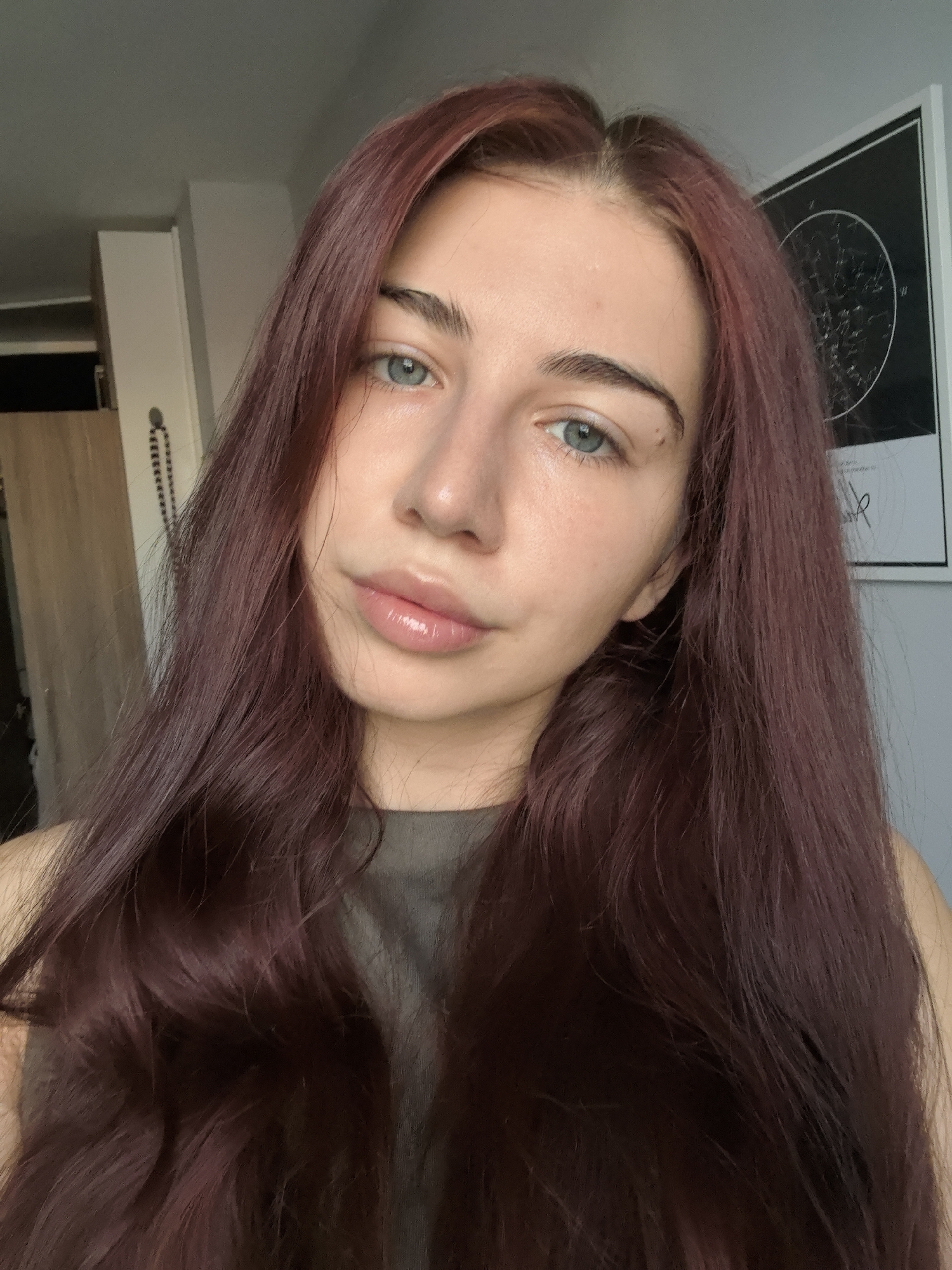 A young woman with long, dark red hair is photographed from the chest up in a selfie. She has blue eyes and full lips with lip gloss. She is wearing a dark grey top. The background shows a wall with a framed star map and a wardrobe with wooden doors. The image is a close-up portrait of the woman. The location is not specified within the image.