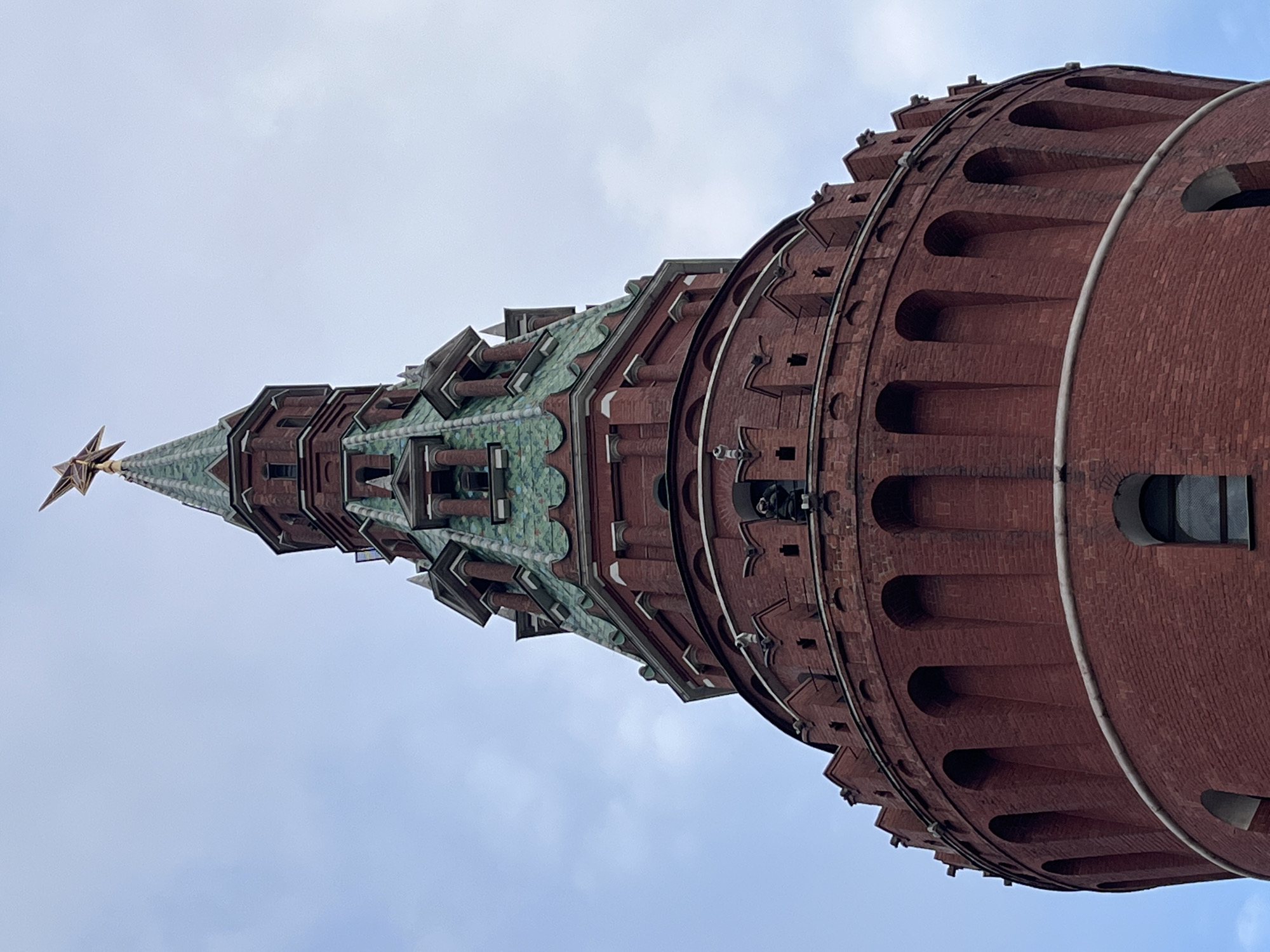 The image shows a low-angle view of the Trinity Tower, part of the Moscow Kremlin in Russia. The tower is constructed of red brick, with a green tiled roof and a gold star at the top. The sky is visible in the background, and the tower is the primary subject.