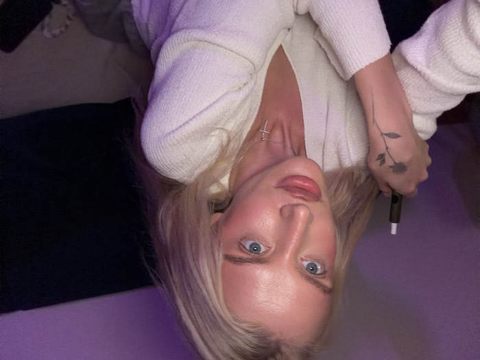 The image captures a close-up, chest-up view of a female individual in an indoor setting. The subject has long blonde hair, blue eyes, and is looking directly at the camera. She exhibits prominent facial features, including full lips. She is wearing a light-colored, likely cream or off-white, textured long-sleeved top with a V-neck. A silver-toned chain necklace with a cross pendant is visible around her neck. Her left hand is positioned near her chest, holding a dark-colored cylindrical vaping device, possibly a heated tobacco product, with a white tip. A black, linear tattoo depicting a rose with a stem and leaves is present on her left forearm/wrist. The background is dimly lit, showing a purple ambient light on the left side and dark, indistinct elements on the right. A light-colored cushion or pillow is partially visible in the lower right corner. This scene is located in Belgrade, Serbia.
