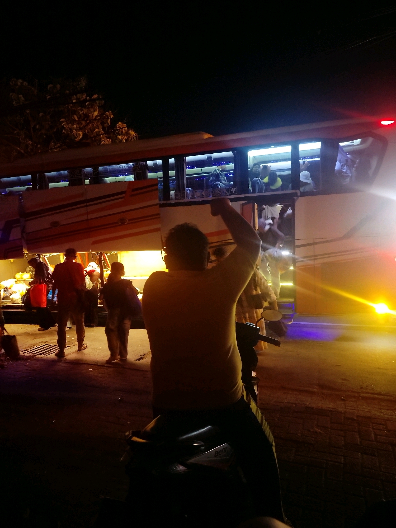 The image depicts a nighttime scene at what appears to be a bus station or roadside stop in Mojokerto, Indonesia. A large bus, illuminated by interior lights and external lighting, is the central focus. Several people are visible, some inside the bus looking out the windows, and others gathered around the bus, appearing to be boarding or disembarking.
In the foreground, a person is seated on a scooter, with their right arm raised in a gesture. Their back is to the camera. The scooter is dark-colored and partially obscured by shadow. The ground is paved with what looks like cobblestones.
To the left of the bus, there are piles of luggage or goods, some in bags and some loose, illuminated by artificial light. A few individuals are standing near this area, and one person in a red shirt is facing away from the camera. Another person in dark clothing is walking away from the bus.
The overall atmosphere is dimly lit, with strong light sources coming from the bus and streetlights, creating harsh shadows. Trees with foliage are visible in the upper left corner against the dark sky. The red taillight of the bus is illuminated. The scene suggests a moment of transit or waiting, possibly at the end of a journey or at the start of a new one, given the presence of luggage and people in a boarding area.