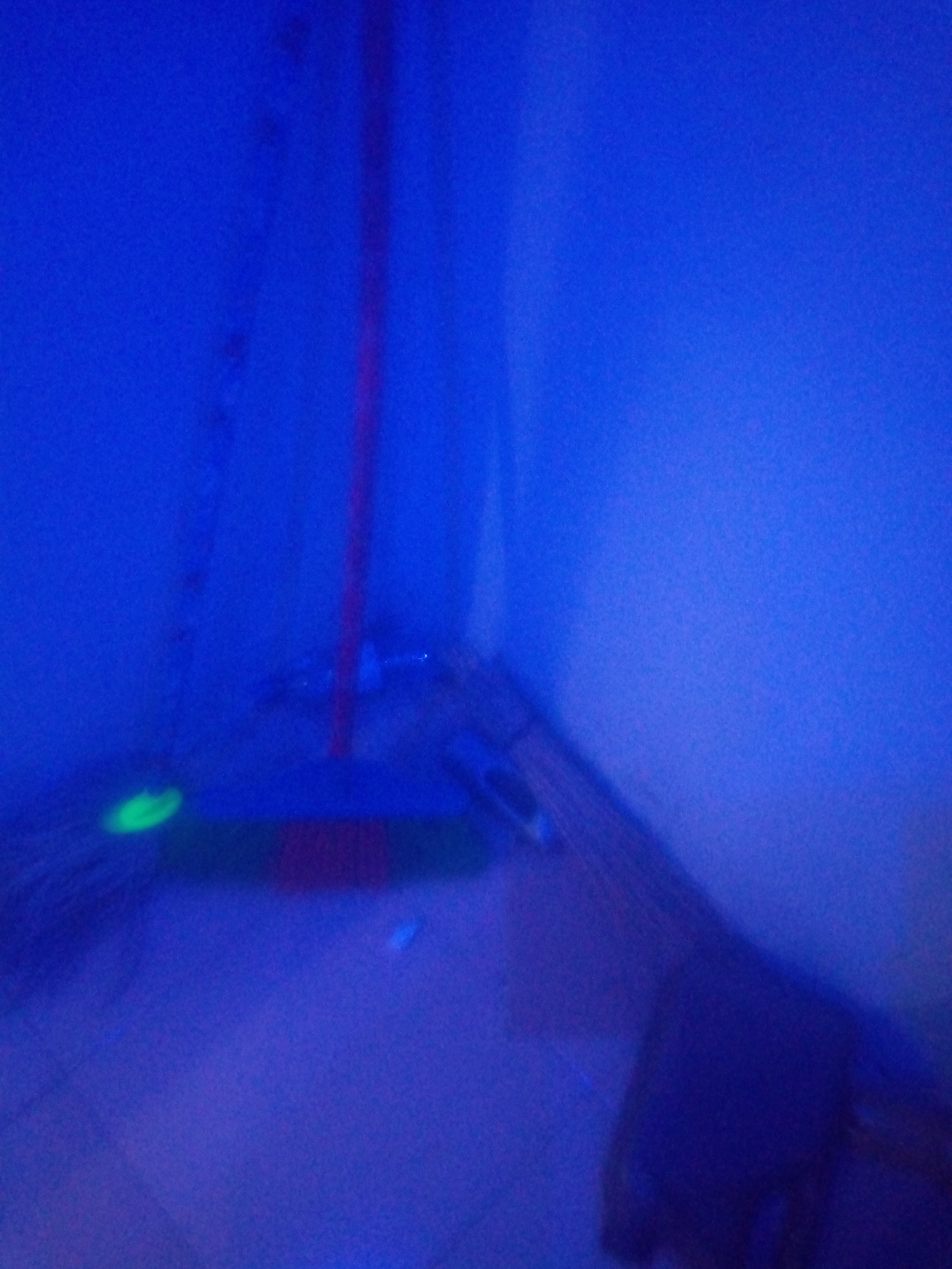The image captures a dimly lit, indoor corner of a room, bathed in a strong blue light, possibly from a blacklight. A red-handled mop is leaning against the wall, its bristles resting on the floor. A faint green glow emanates from the bristles of the mop, suggesting the presence of a fluorescent substance. The floor appears to be tiled, with the tiles reflecting the blue light. The walls are a solid, unadorned blue. The overall impression is one of a cleaning in progress or recently completed, with an unusual light source adding a mysterious quality to the scene. The location in Jalingo, Nigeria, is not explicitly indicated within the image itself.