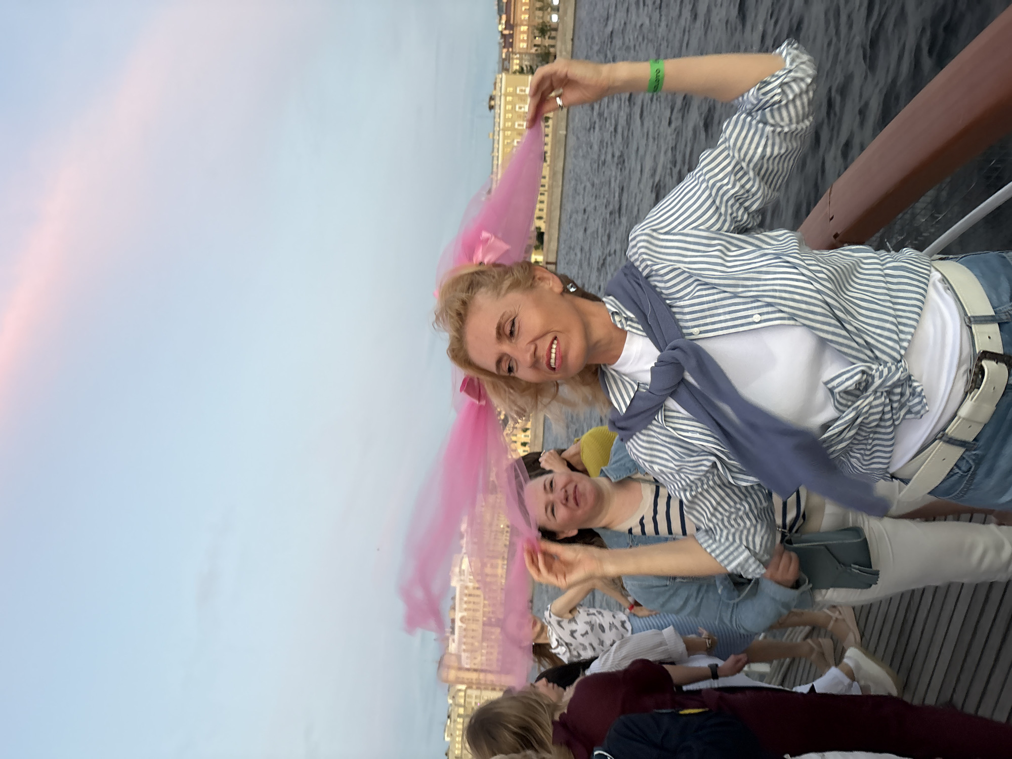 A group of people are on a boat trip on a river in Saint Petersburg, Russia, at dusk. The sky is light blue with some pink clouds. The people are dressed casually. The woman in the foreground is wearing a striped shirt and jeans. She is holding a pink veil. The other people in the image are blurry and out of focus. The background shows buildings on the riverbank. The water is dark and choppy.