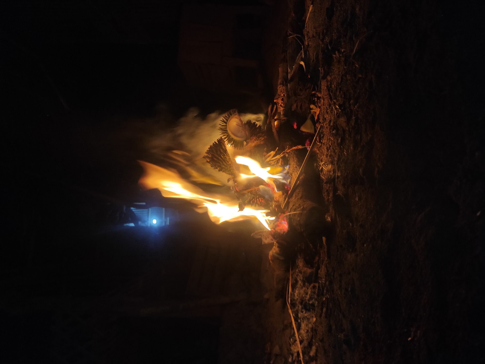 The image shows a nighttime scene of a small bonfire in what appears to be an outdoor, perhaps rural or less developed area, possibly on the outskirts of Lhokseumawe, Indonesia. The fire is the central subject, with bright orange and yellow flames rising against a dark background. The fire is built on what looks like a patch of dry earth or dirt, with scattered twigs, leaves, and some larger pieces of wood. Two spiky objects, possibly durian husks or similar natural material, are positioned within the flames, contributing to the fire's intensity. Smoke billows upwards from the fire. In the background, there is a dimly lit structure or area with a distinct blue light source visible, suggesting it might be a distant building or artificial light. The overall impression is one of a rustic, perhaps temporary, outdoor gathering or activity occurring in darkness, illuminated primarily by the fire.