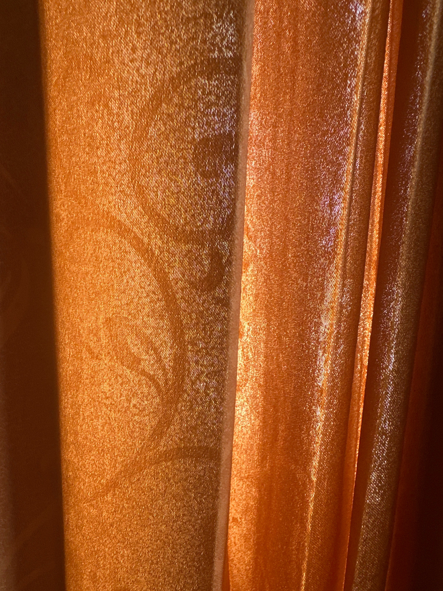 The image is a close-up, eye-level, horizontal shot of orange fabric. The fabric is draped, creating vertical folds and subtle variations in light and shadow. The texture of the fabric appears to be woven, with a slight sheen that reflects the light. There is a faint, swirling pattern visible on the fabric on the left side of the image. The lighting suggests a warm, possibly artificial light source. The image does not contain any recognizable landmarks or features that would indicate a specific location in Margamulya, Indonesia. There are no people, objects, actions, or interactions depicted.