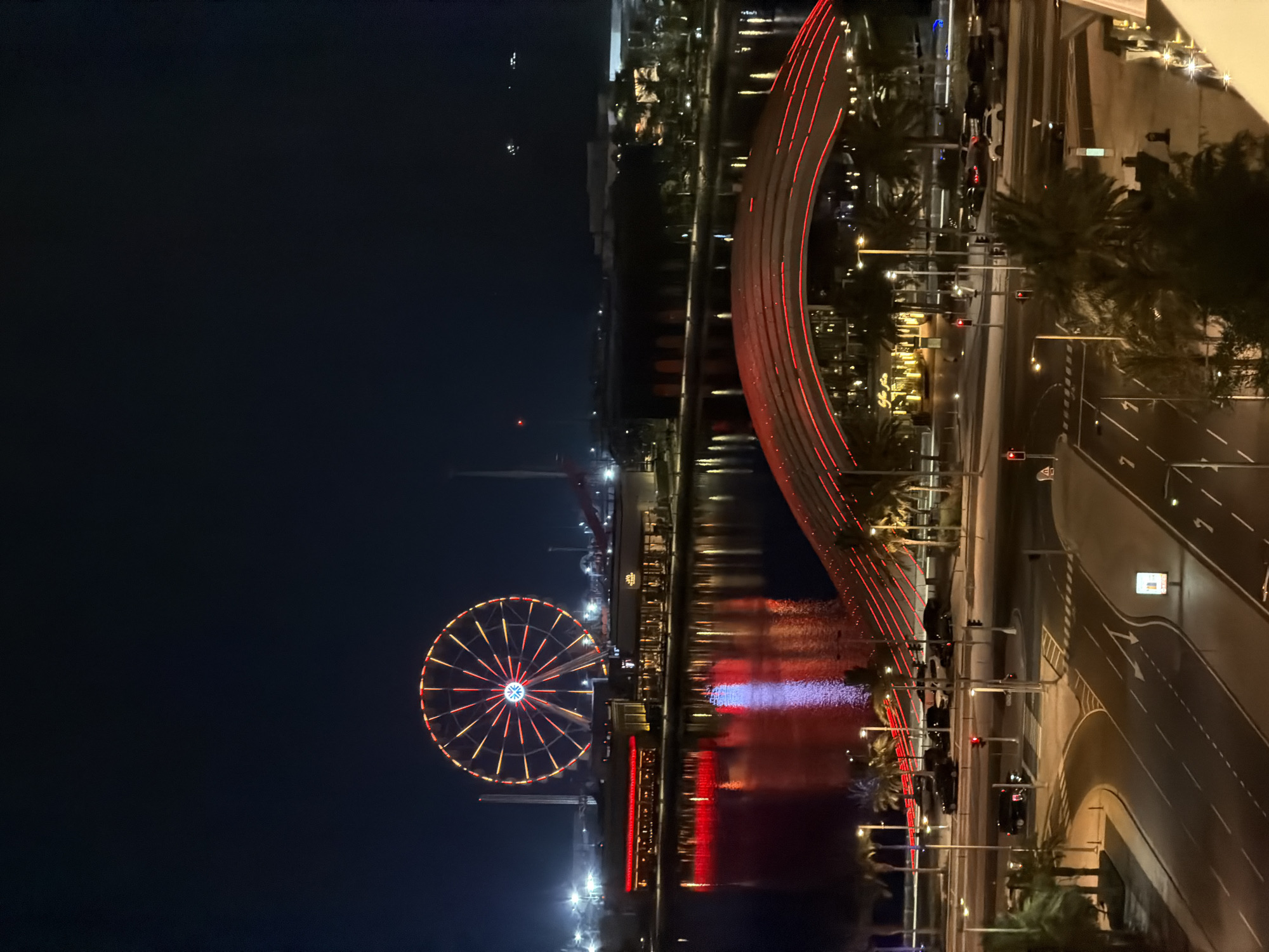 This media file captures a nighttime scene in an urban waterfront area, likely a tourist or entertainment district. The image is oriented vertically, with the night sky taking up the left portion and the illuminated cityscape occupying the right.
The prominent subjects are a large Ferris wheel, lit with red and yellow lights in a spoke pattern, and a distinctive curved bridge or walkway, also illuminated with red lights. The bridge's reflection is visible on the water below. The water also reflects other lights, including areas of purple and red. Beyond the water and bridge, there's a densely lit area with buildings, palm trees, and other structures. A street with traffic lights, road markings, and parked cars runs along the right side of the frame, with streetlights and traffic signals visible.
The time of day is clearly night, with the dark sky and artificial lighting dominating the scene. The weather appears to be clear, allowing for reflections on the water. There are no visible people in the frame, but the presence of cars and the illuminated Ferris wheel suggest a place that is active or popular. The overall atmosphere is vibrant and festive due to the extensive use of colorful lighting. The location appears to be a modern city, possibly a port or a developed waterfront with leisure facilities.
No discernible text is visible in the image.