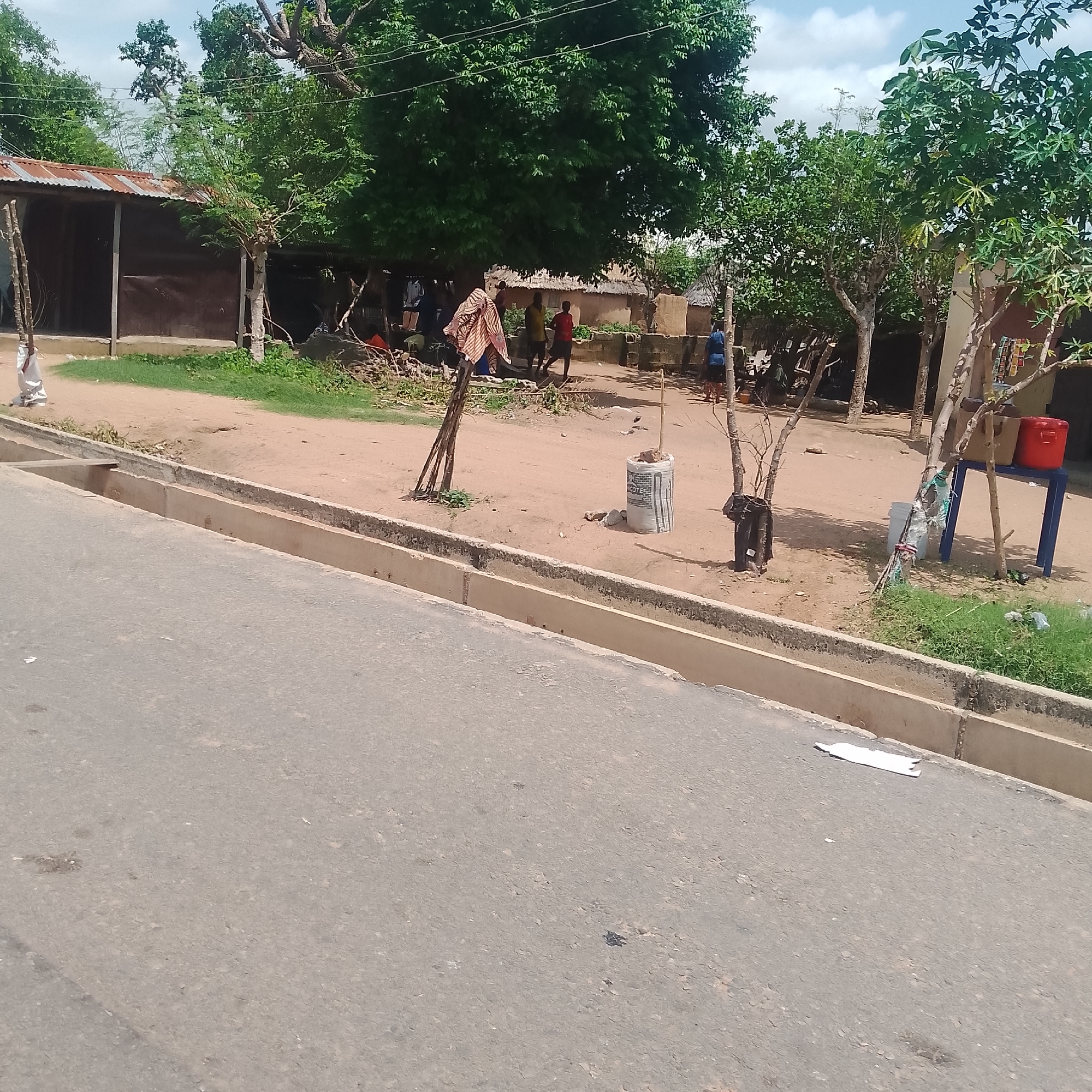 The image depicts a street scene in Jalingo, Nigeria. In the foreground is an asphalt road with a concrete gutter. Beyond the gutter is a dirt path with sparse vegetation. On the left, a corrugated metal-roofed structure with dark brown walls stands partially visible. Several people are present in the background, some standing near the structure and others further back near mud-brick buildings. A blue table with two red containers and a brown box is visible to the right. Various objects are scattered around the area, including a white cylindrical bag with text, branches, and trash. The environment appears to be a semi-urban or rural setting under a cloudy sky.
