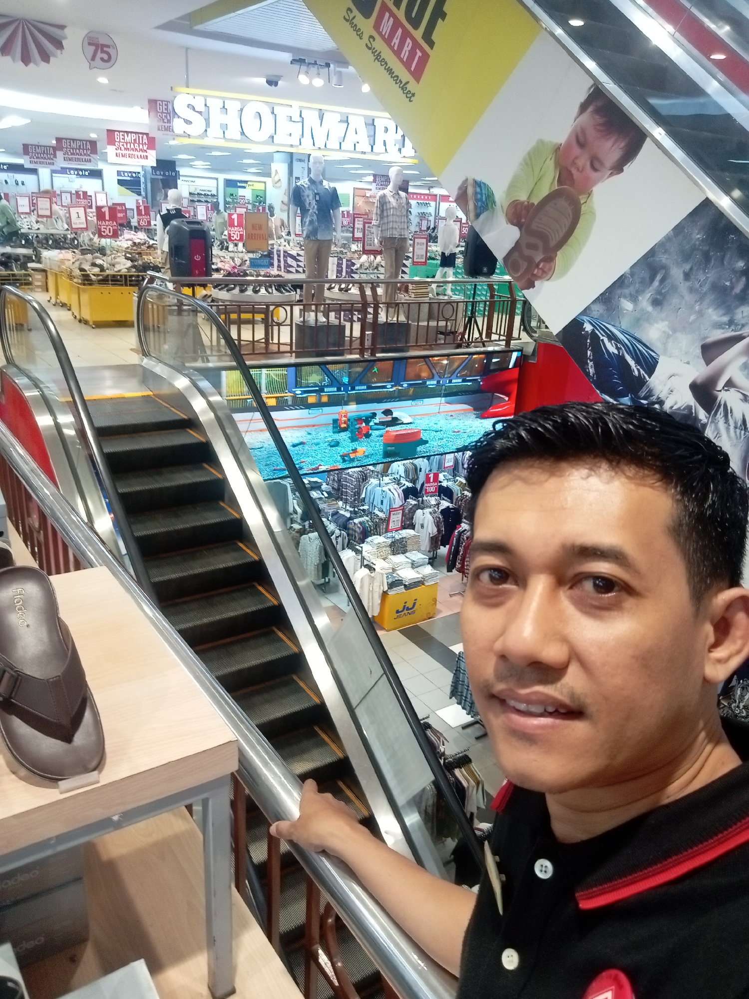 A selfie taken inside a shopping mall in Batam, Indonesia, features a man in the foreground with a bright red and black collared shirt. Behind him, a department store called "Shoemart" is visible, displaying shoes and clothing on racks and shelves. An escalator with metal railings and black steps is prominently featured. A large advertisement for a baby holding a shoe is affixed to the side of the escalator. The mall appears to be busy, with other shoppers and displays visible in the background. The man's expression is neutral, and he is holding onto the escalator railing with his right hand. The lighting in the mall is typical of an indoor retail environment, with fluorescent lights illuminating the displays. The overall impression is of a typical shopping experience in a modern mall.