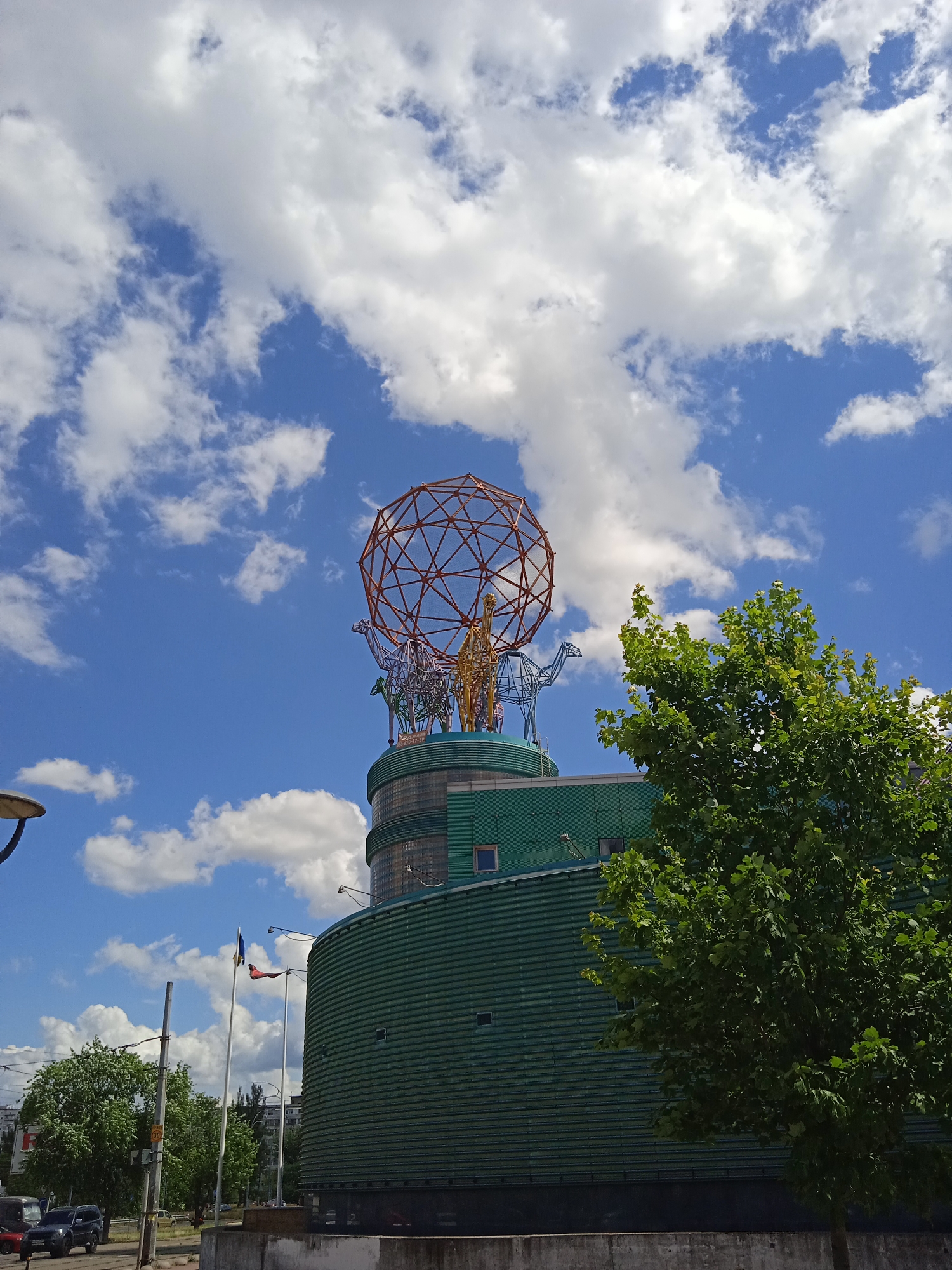 A low-angle outdoor shot shows a modern, green, cylindrical building with a large, orange, spherical, geodesic dome-like sculpture on its roof. The sculpture is supported by several stylized animal figures, including what appear to be giraffes. The building has a textured, ribbed facade and several small, square windows. In the foreground to the right, a large, leafy green tree partially obscures the building. The background features a bright blue sky with scattered white clouds. To the left of the building, a street is visible with several cars parked and driving. Two flagpoles with flags are also present to the left of the building. The overall scene suggests an urban environment under a bright, partly cloudy sky.