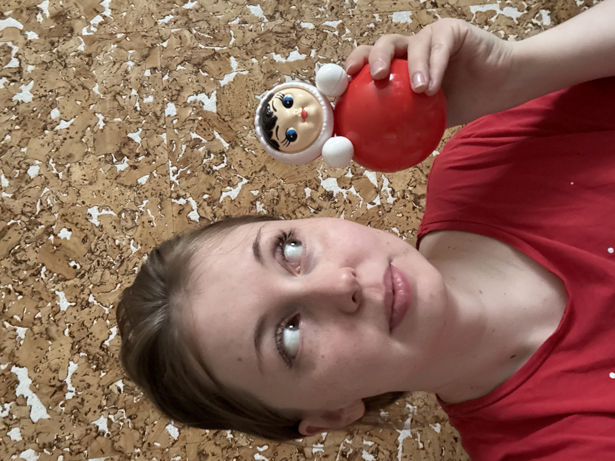 A young woman in a red shirt is holding a red roly-poly toy with a white-hooded face. The background appears to be a corkboard wall.